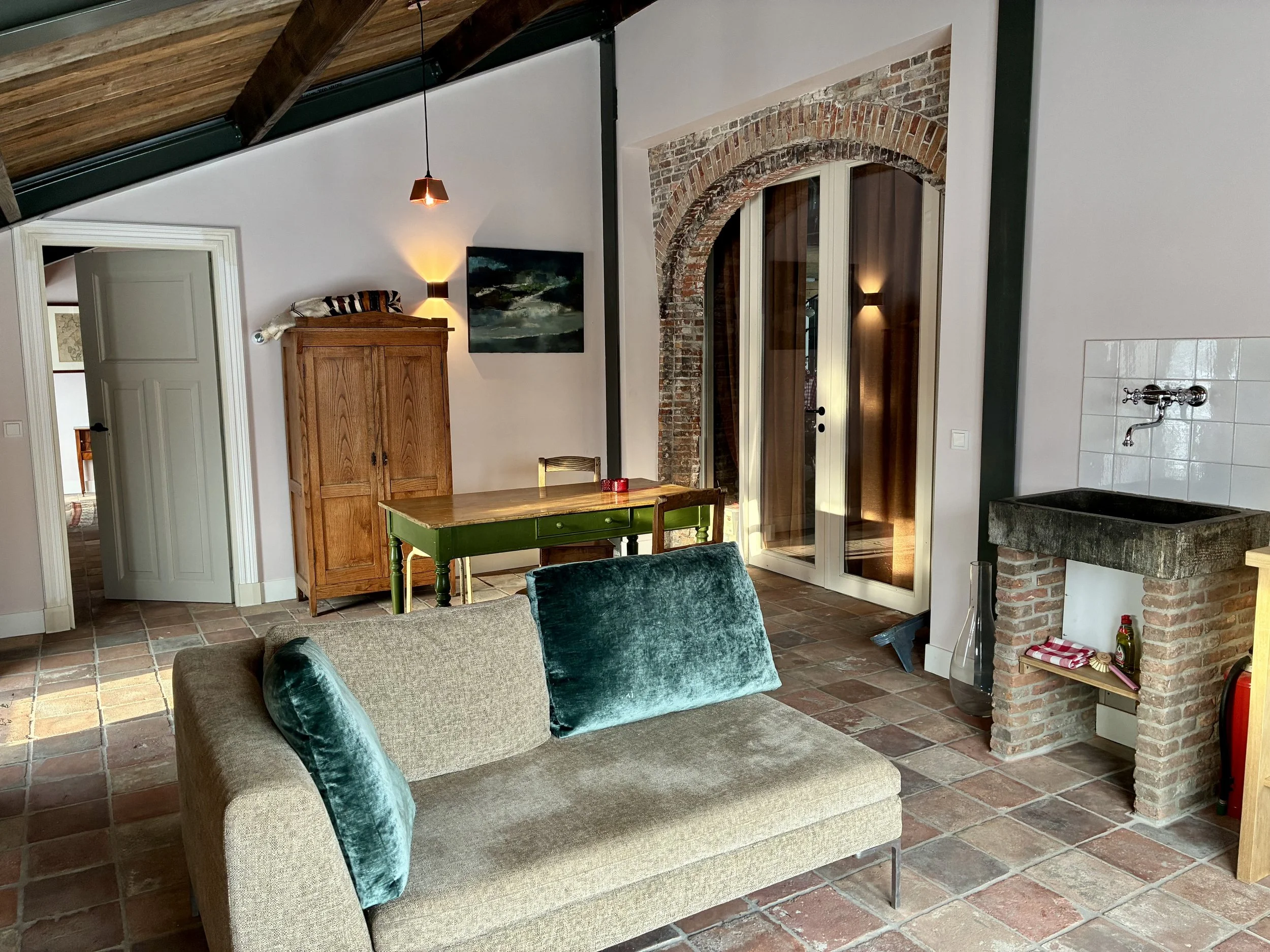 Cozy living room with a beige sofa and green pillows, wooden furniture, exposed brick arch doorway, tile flooring, and a sink with wall-mounted faucet.