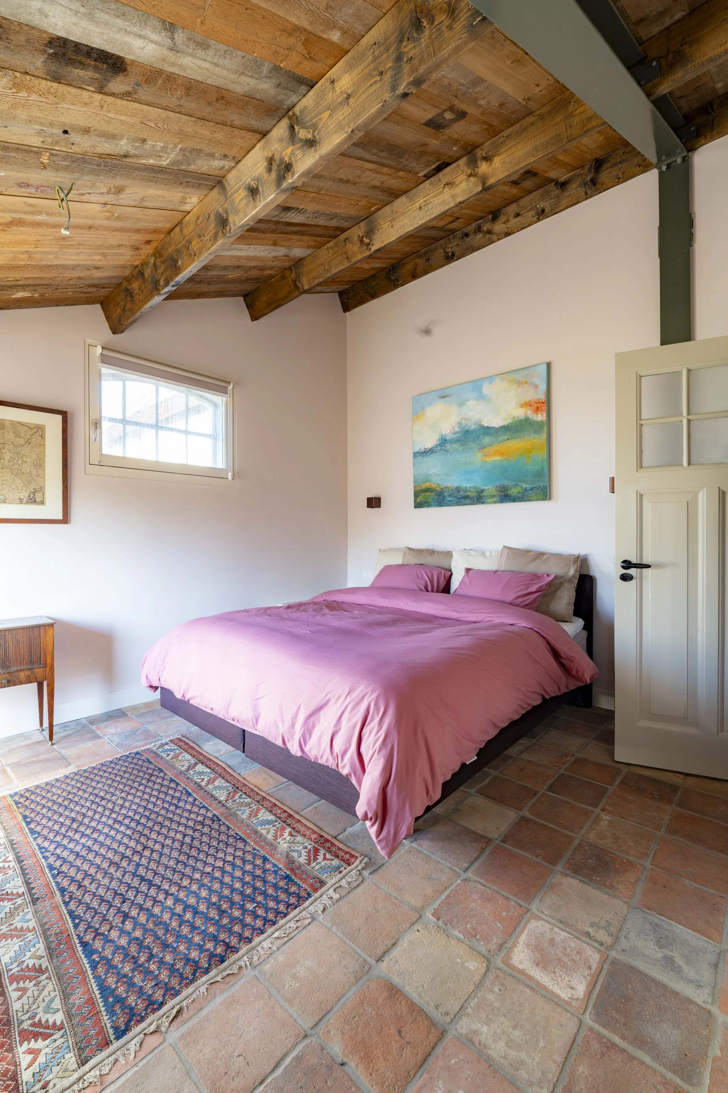 A bedroom with a wooden ceiling and beams, a pink bed with beige pillows, a small wooden nightstand, a colorful area rug, a window, and a door. There is a landscape painting on the wall.