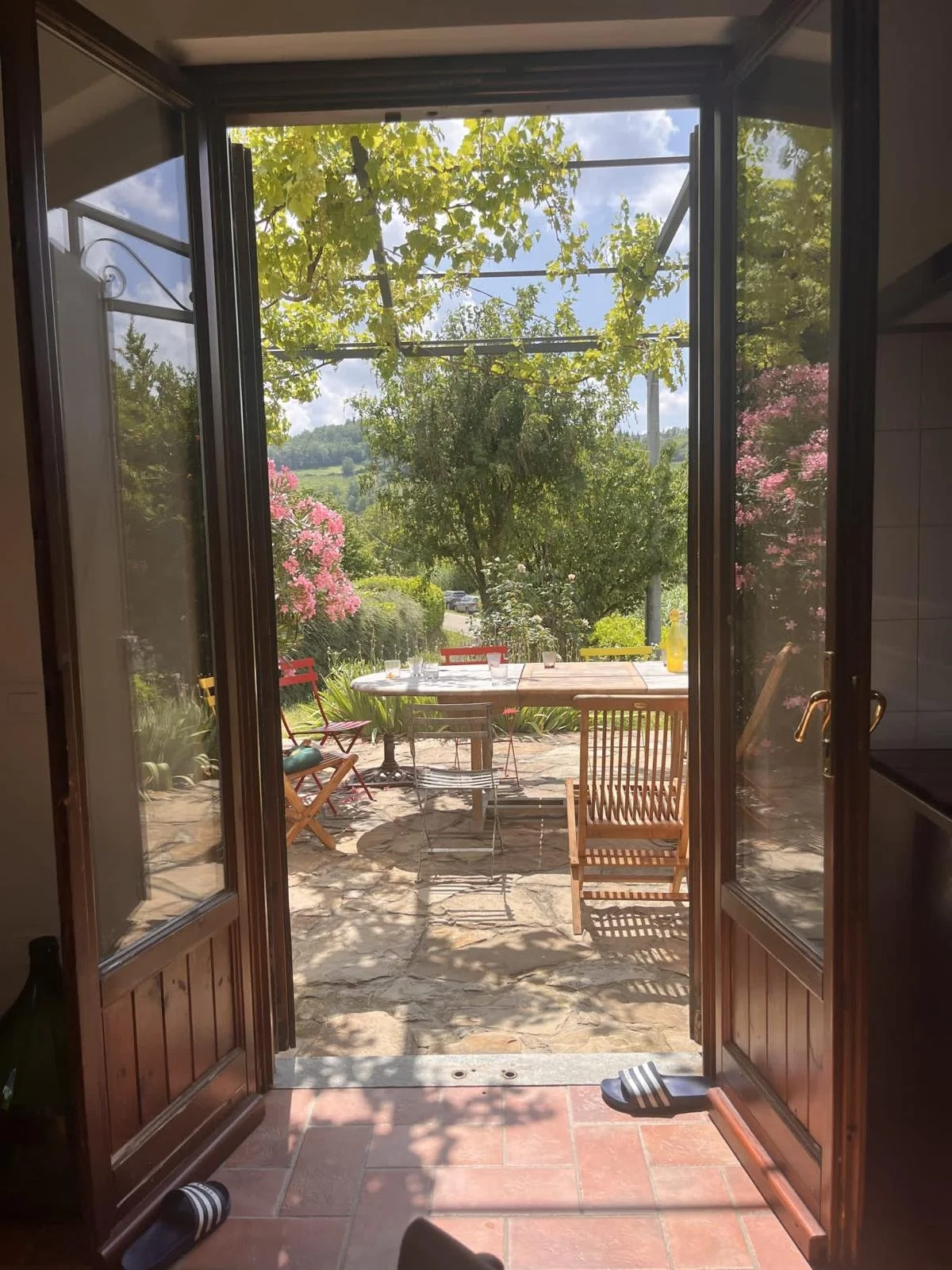 View through a doorway to a patio with a table and chairs, surrounded by greenery and flowering bushes, under a bright blue sky with some clouds.