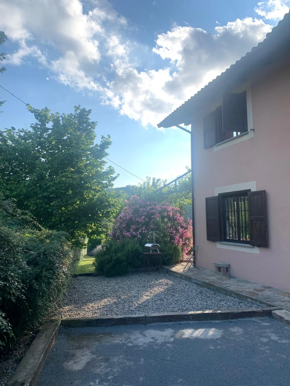 A house with pink exterior walls and dark shutters, a gravel pathway, lush green trees, a flowering bush with pink blossoms, outdoor furniture, and a partly cloudy sky.