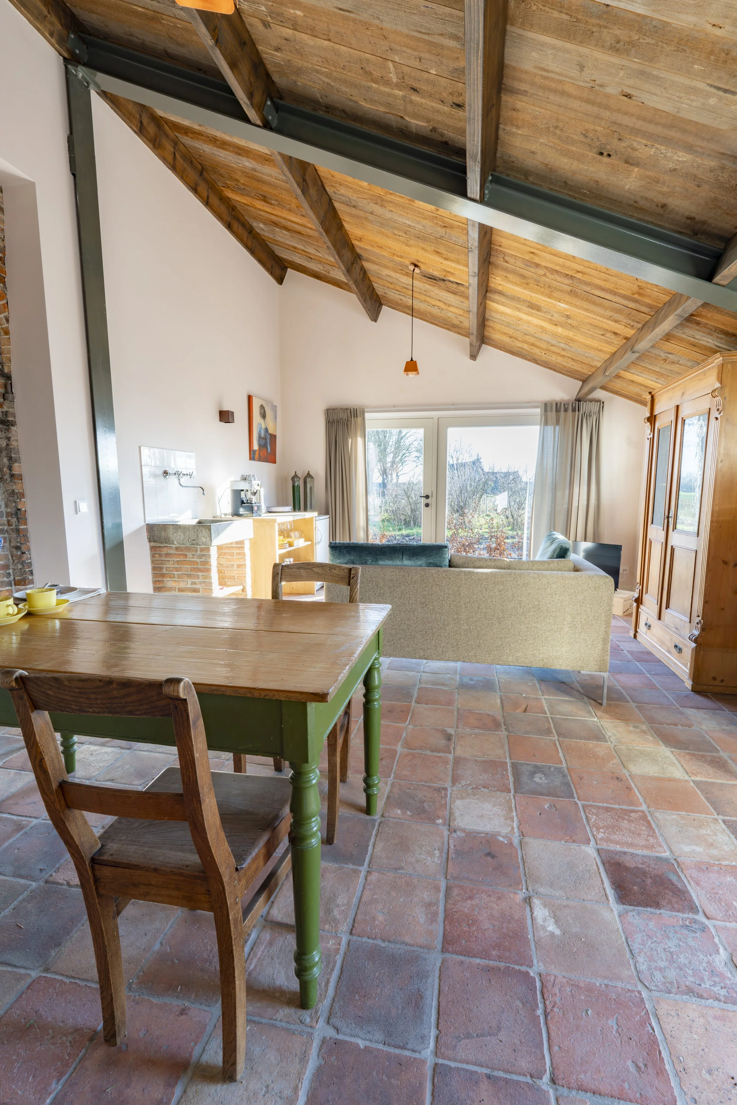 Interior of a living room with exposed wooden ceiling beams, a tan sofa, sliding glass doors with curtains, a kitchen area with a countertop, a wooden dining table with a chair, and a wooden armoire, all with terracotta tile flooring.