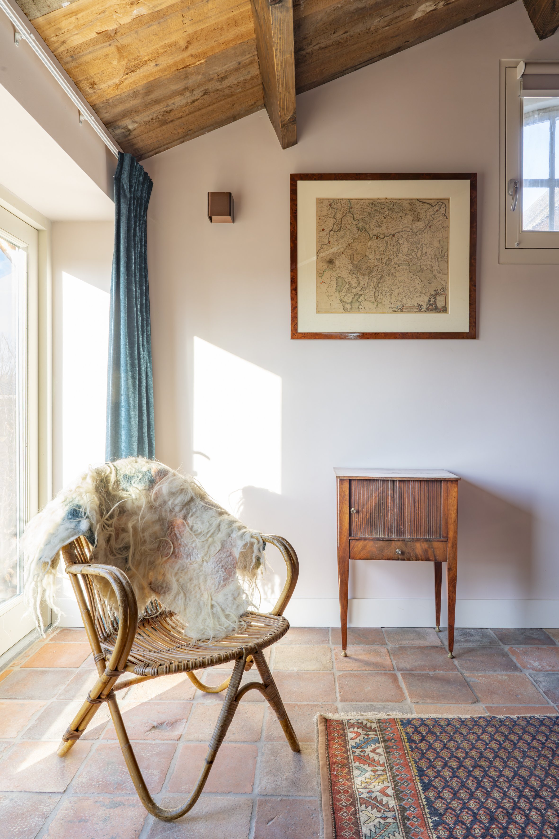 Sunlit room with a wicker chair draped with a wool blanket, a small wooden cabinet, framed map on the wall, and a window with blue curtains.