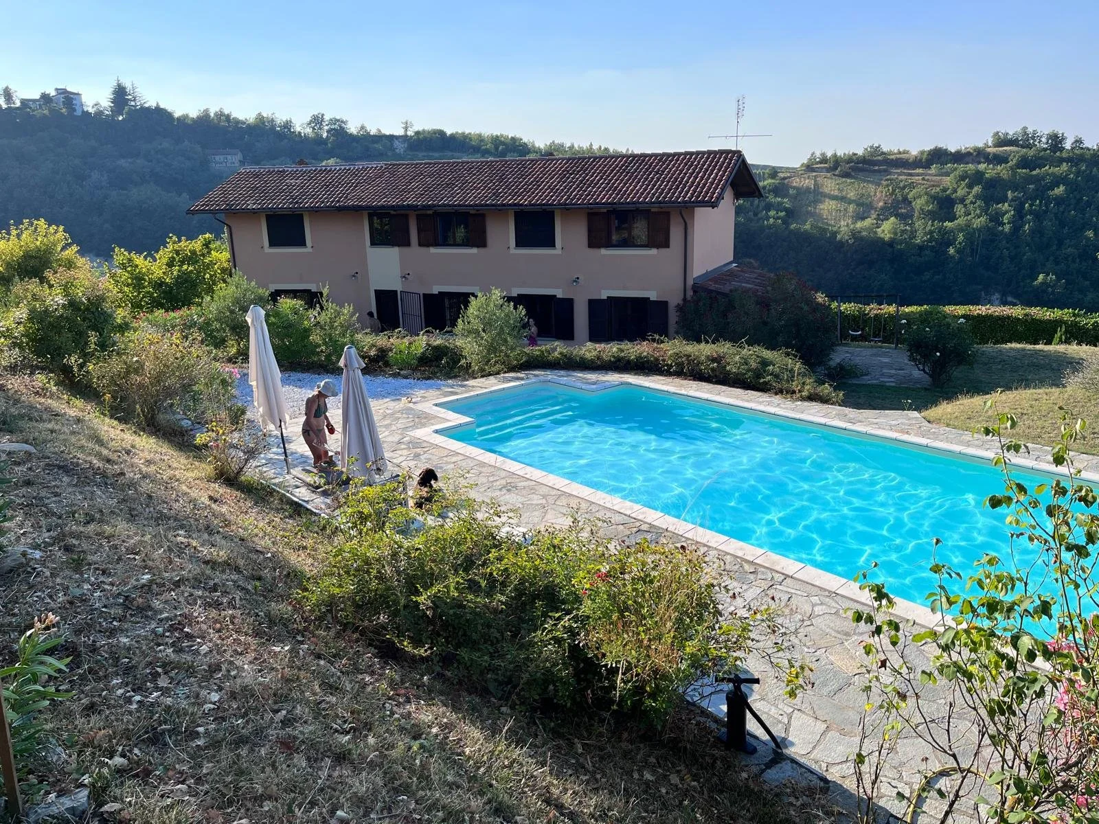 Swimming pool with clear blue water surrounded by a stone deck, adjacent to a two-story house with beige walls and brown shutters, set in a hilly landscape with trees and greenery.