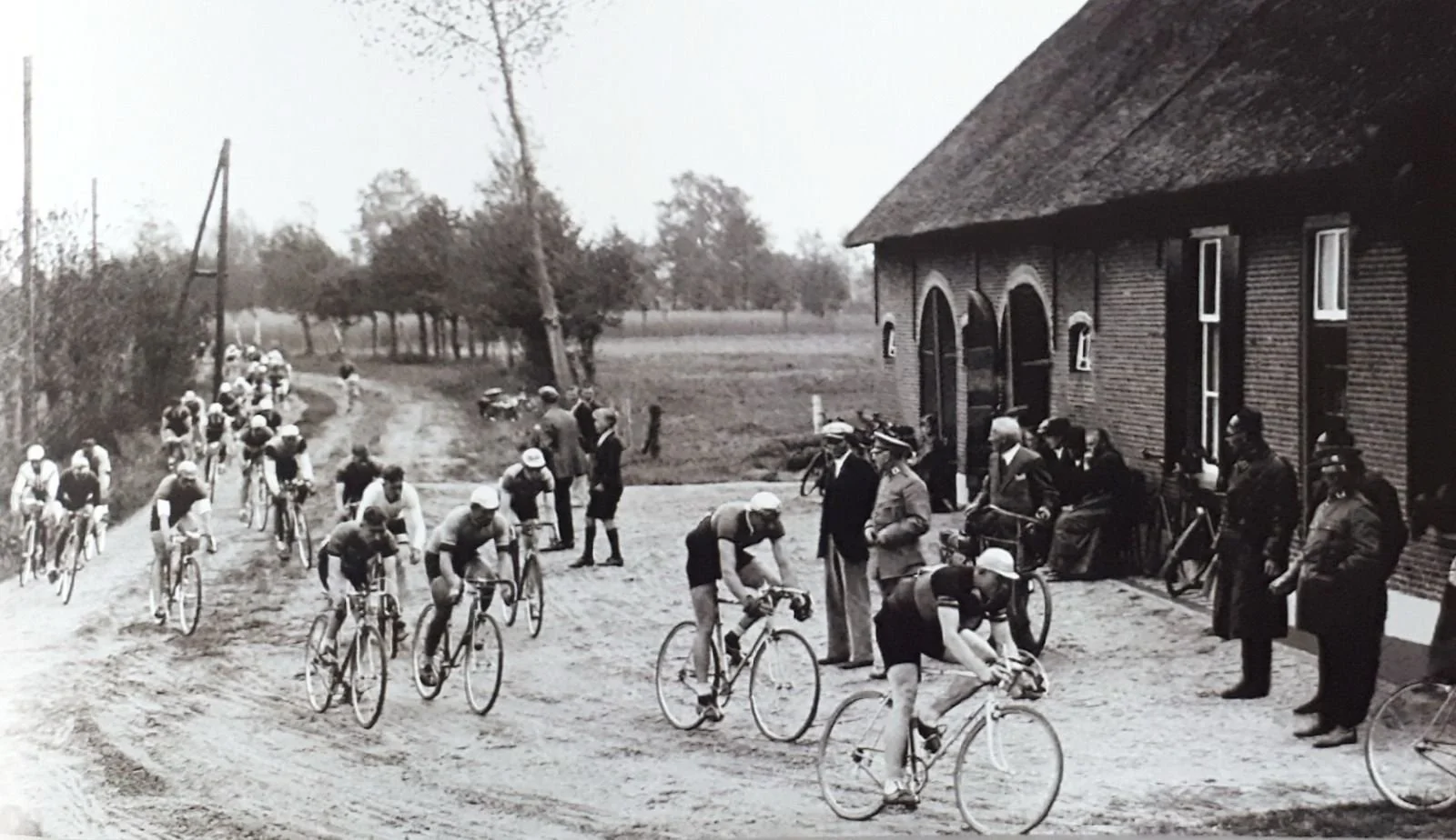 Black and white photo of a cycling race with numerous cyclists passing by a brick house and spectators on a dirt road in a rural area, with trees in the background.