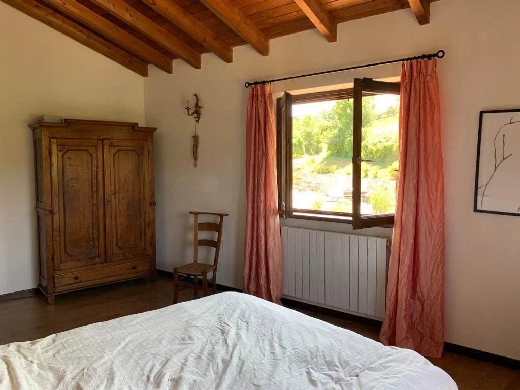 Bedroom with wooden ceiling beams, a wooden wardrobe, a wooden chair, open window with red curtains, a view of trees outside, part of a bed with light-colored sheets, and wall art.