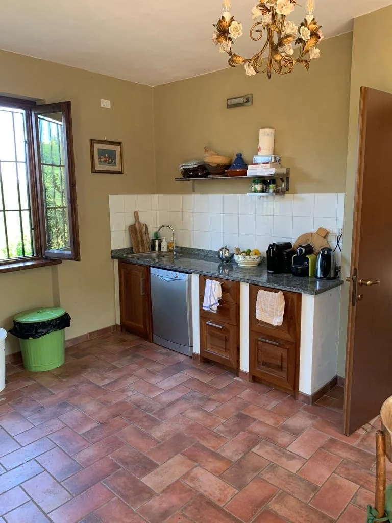 Small kitchen with a window, wooden cabinets, a dark granite countertop, white tile backsplash, and various kitchen appliances on the counter. There are shelves with books and decorative items, a chandelier hanging from the ceiling, and a tile floor.