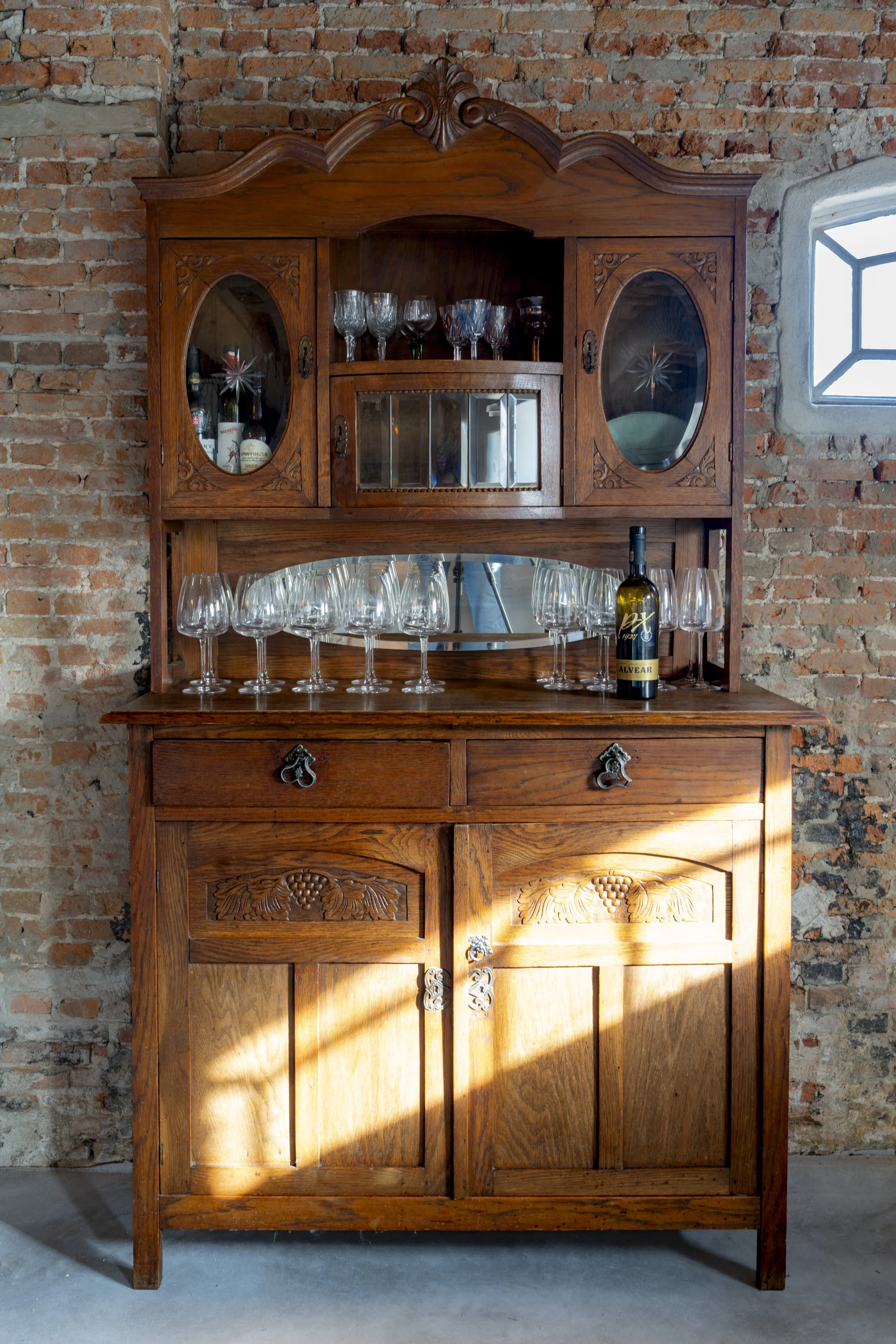 A vintage wooden china cabinet with glass doors and shelves, displaying glassware and wine bottles, set against a brick wall with a small window.