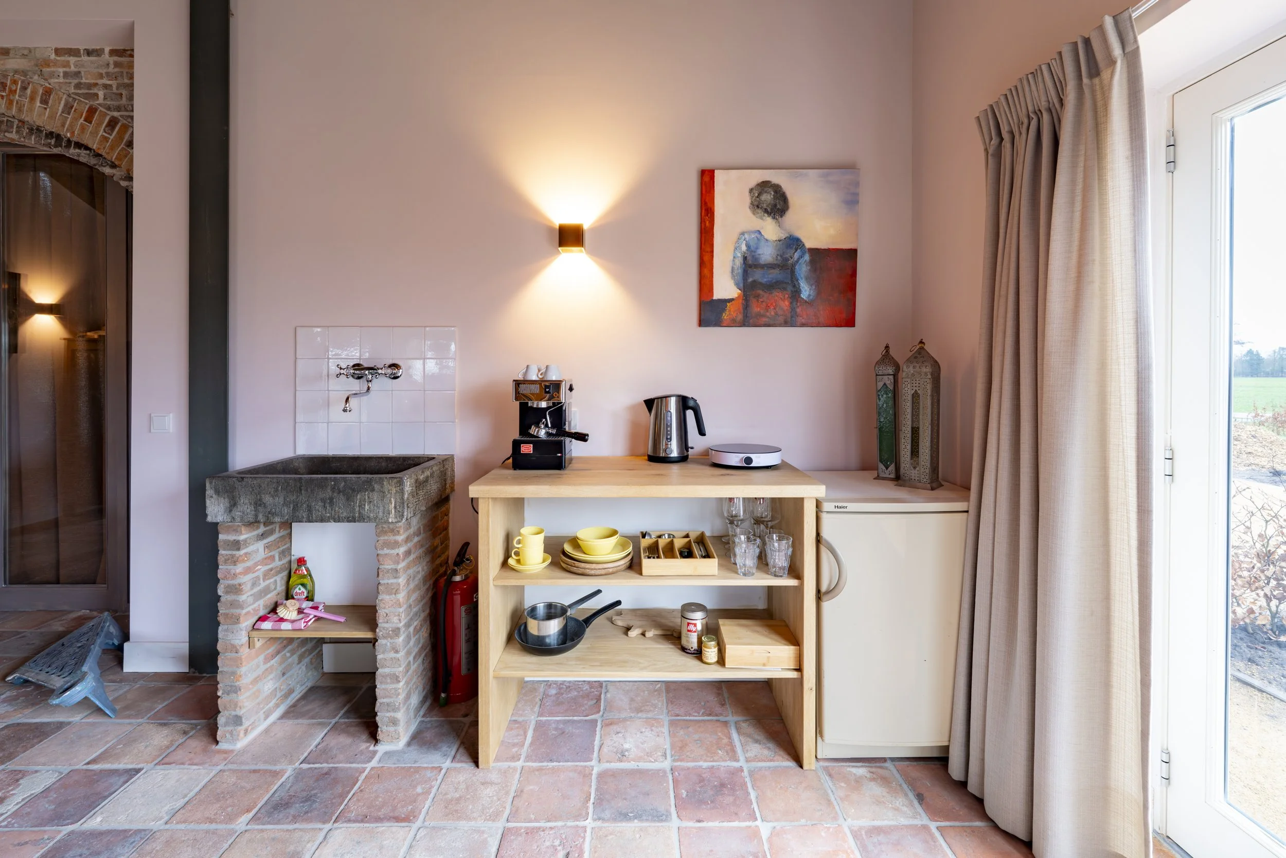 Interior of a rustic kitchen corner with a brick sink, a small wooden shelf with dishes, glasses, pots, and kitchen appliances, a white fridge, artwork of a woman on the wall, and beige curtains covering a glass door.