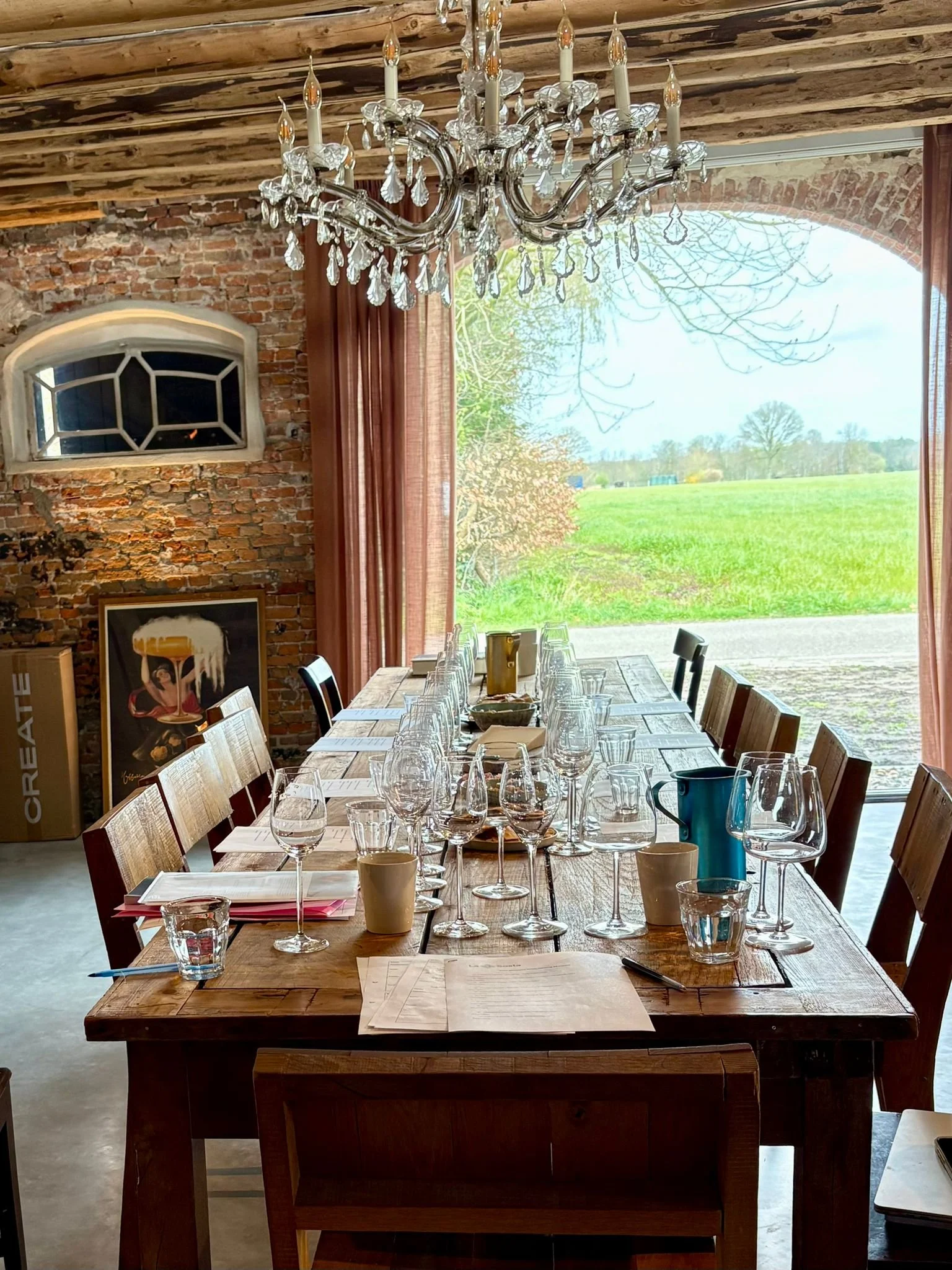 Open-concept living and dining area with rustic wood ceiling and exposed brick wall, featuring a dining table with chairs, a chandelier, a sofa, and a kitchen with modern appliances, large windows showing greenery outside.