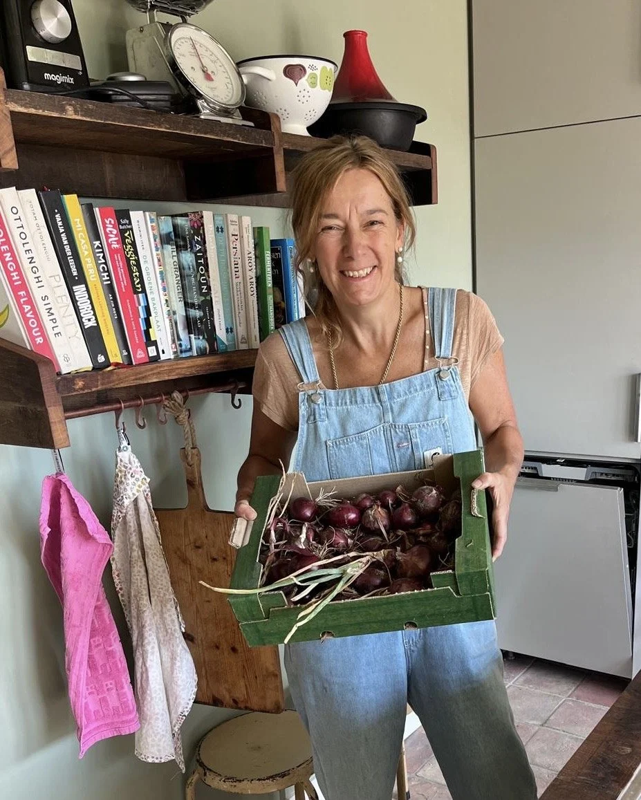 A woman wearing a denim apron and a beige t-shirt, smiling and holding a green crate filled with red onions inside a kitchen. Behind her are books, kitchen utensils, and a white dish rack.