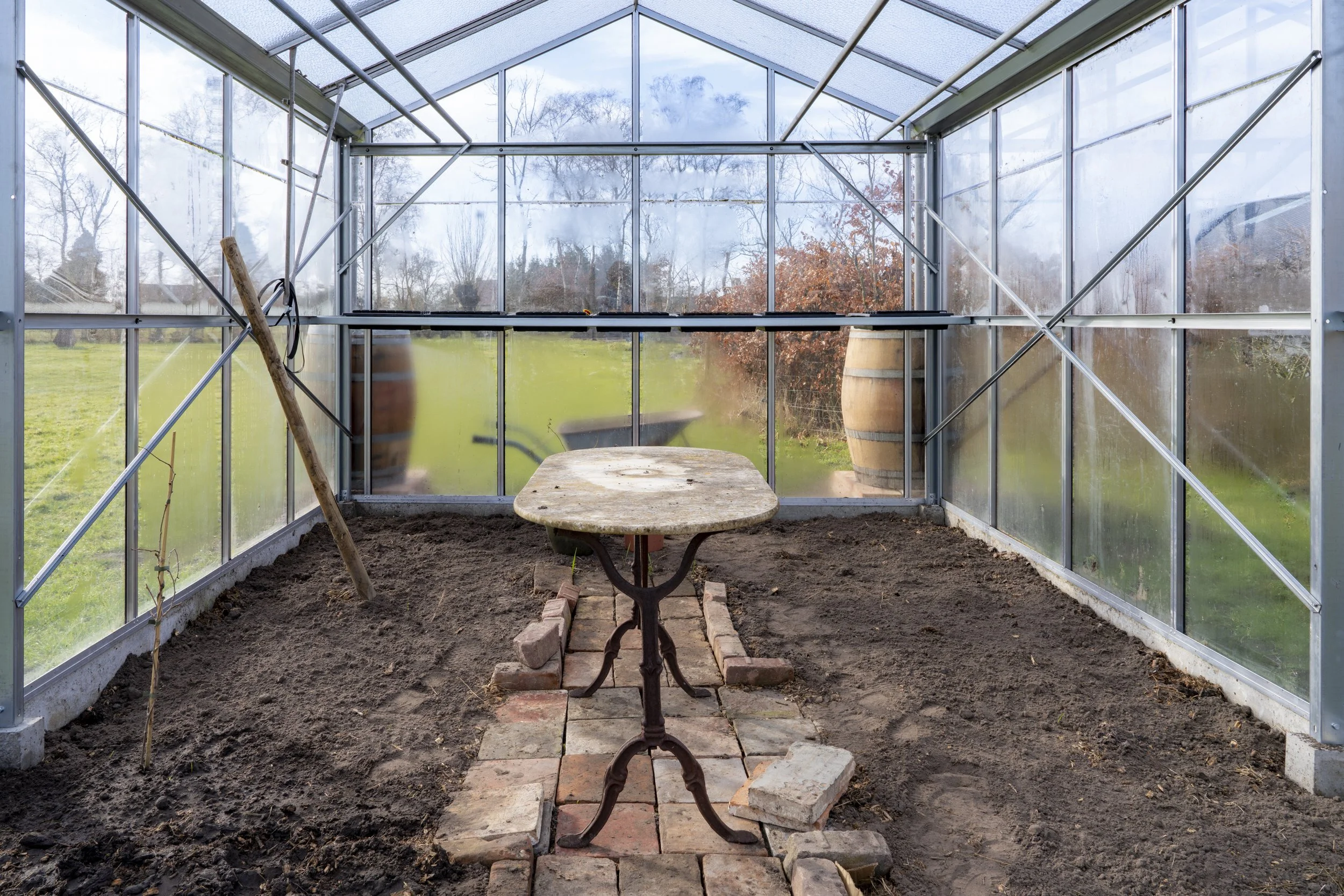 Inside a greenhouse with dirt floor, a wooden table in the center, and a brick pathway leading to the table. There is a wooden stick with a black wire leaning against the left wall, and large barrels outside the greenhouse visible through the glass walls.