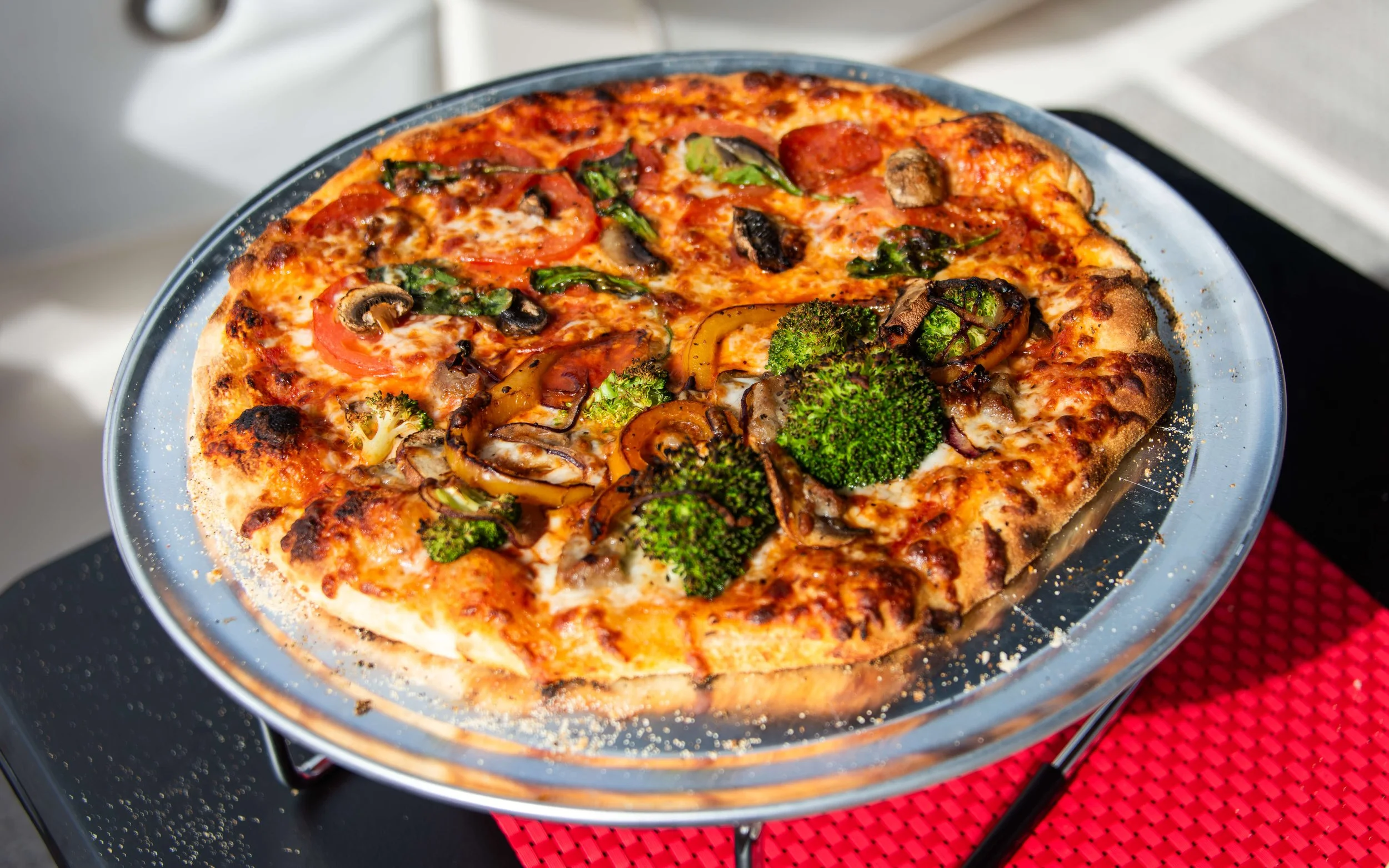 A vegetable pizza with broccoli, mushrooms, tomatoes, and peppers on a crust, on a glass serving plate.