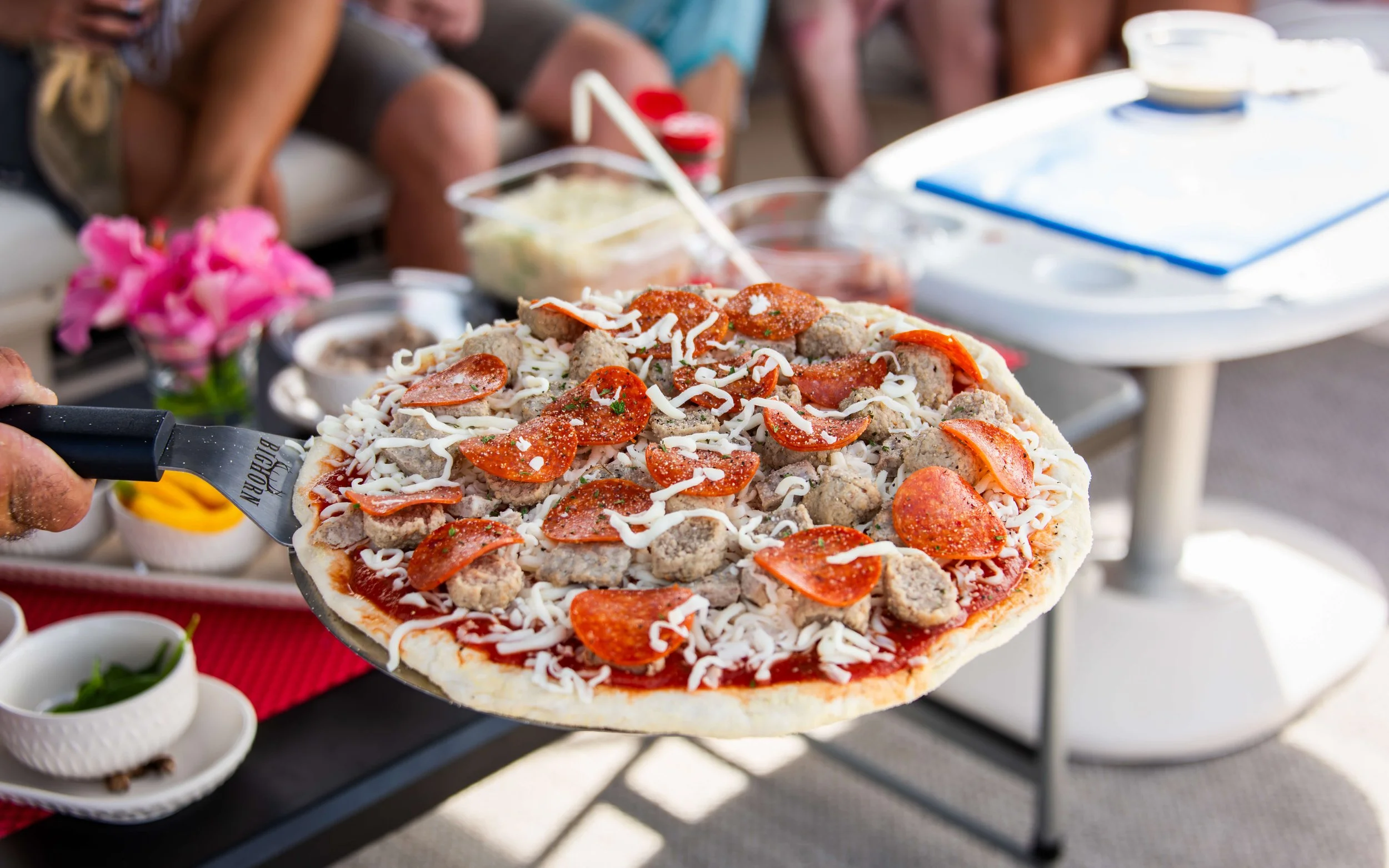 Person holding a pizza with pepperoni, sausage, and cheese at an outdoor gathering.