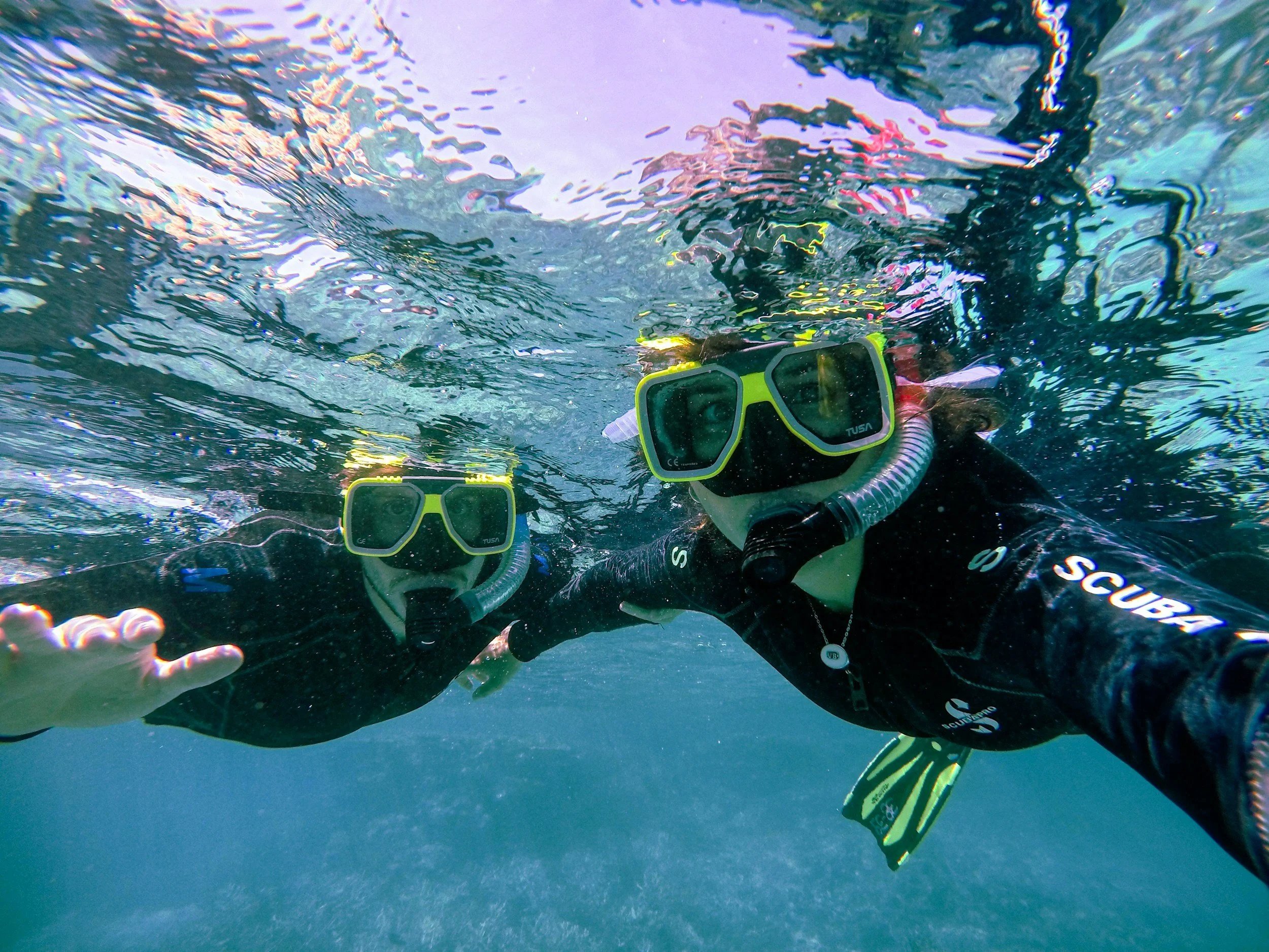Two people wearing scuba gear underwater taking a selfie. They are wearing masks, snorkels, and wetsuits.