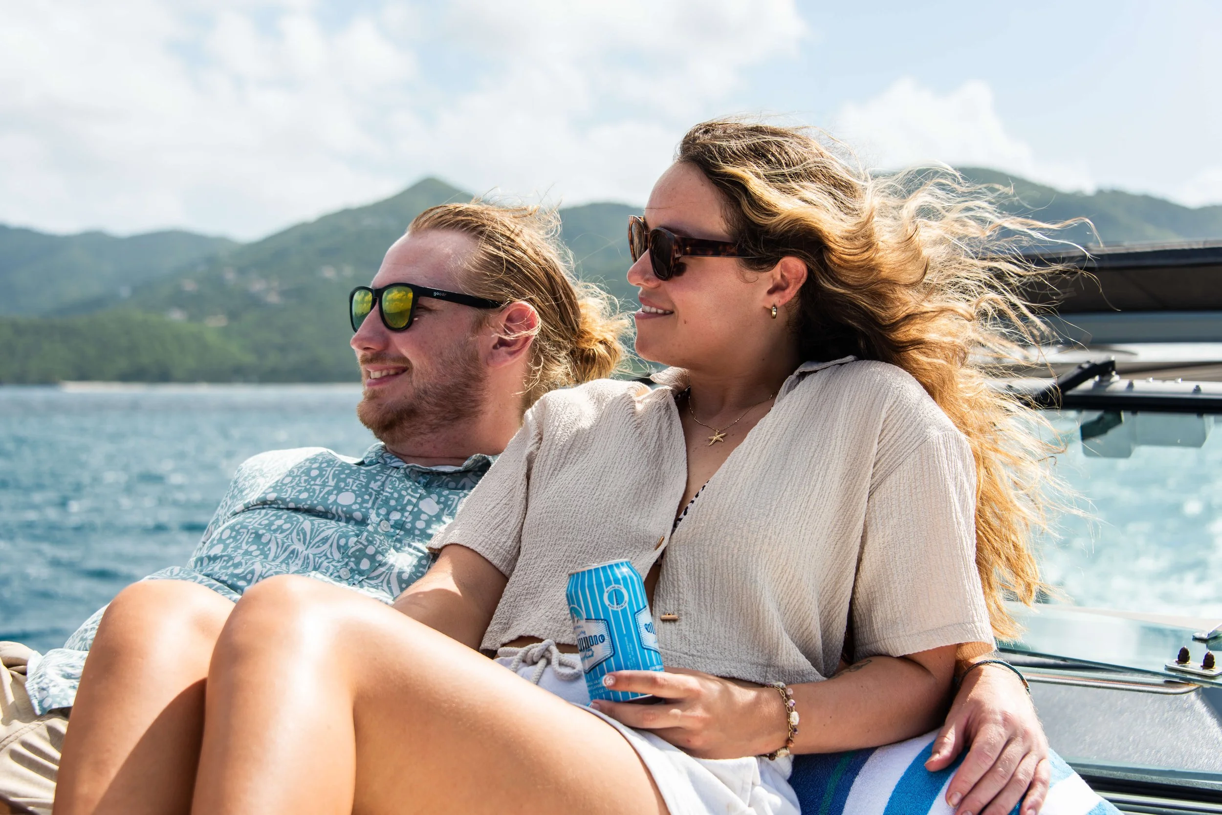 Two people relaxing on a boat, smiling, with mountains and water in the background, and holding a can of drink.