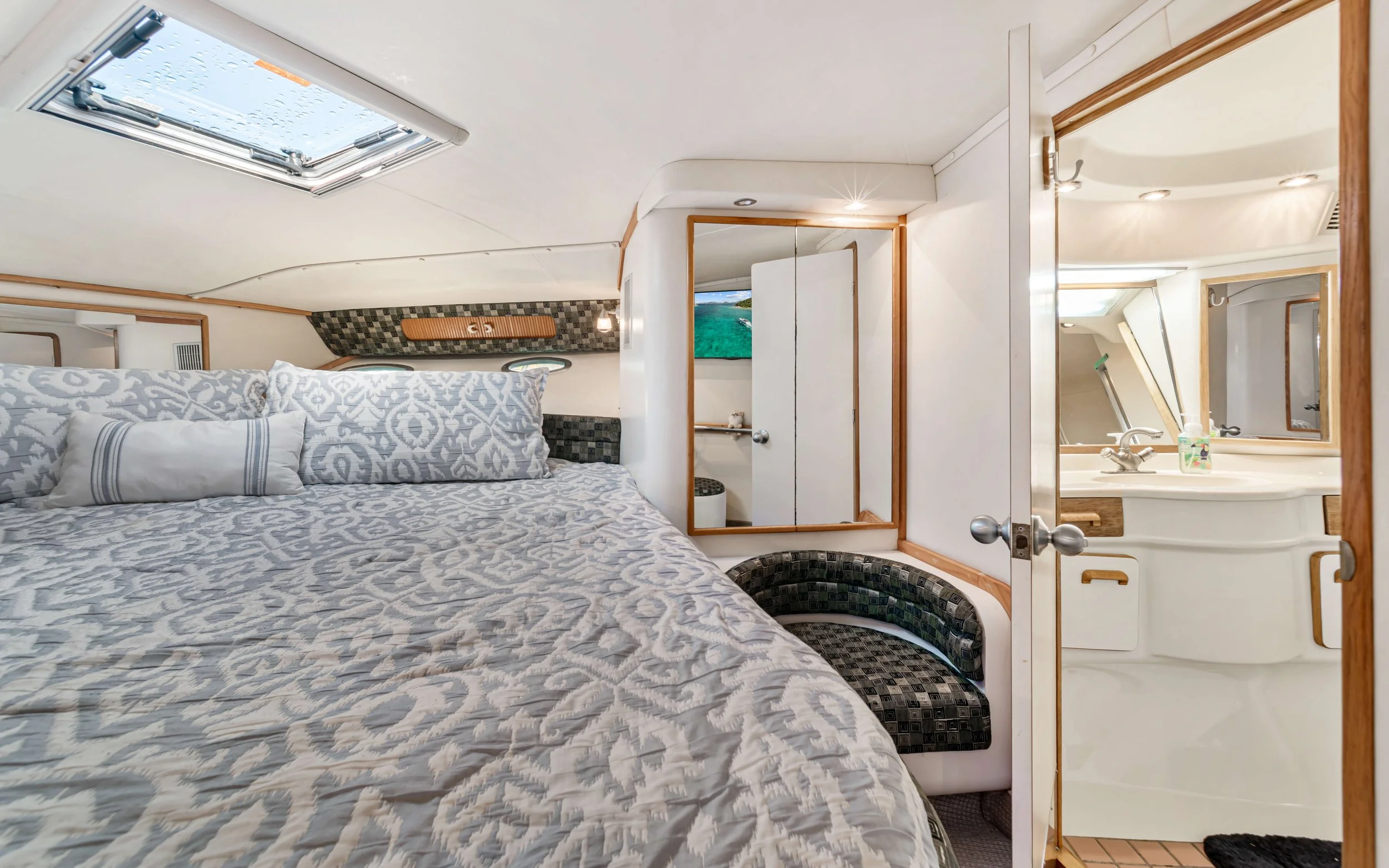 Interior of a compact bedroom with a bed covered in patterned bedding, small pillows, and a window and skylight overhead, adjacent to a bathroom with a sink and mirror.