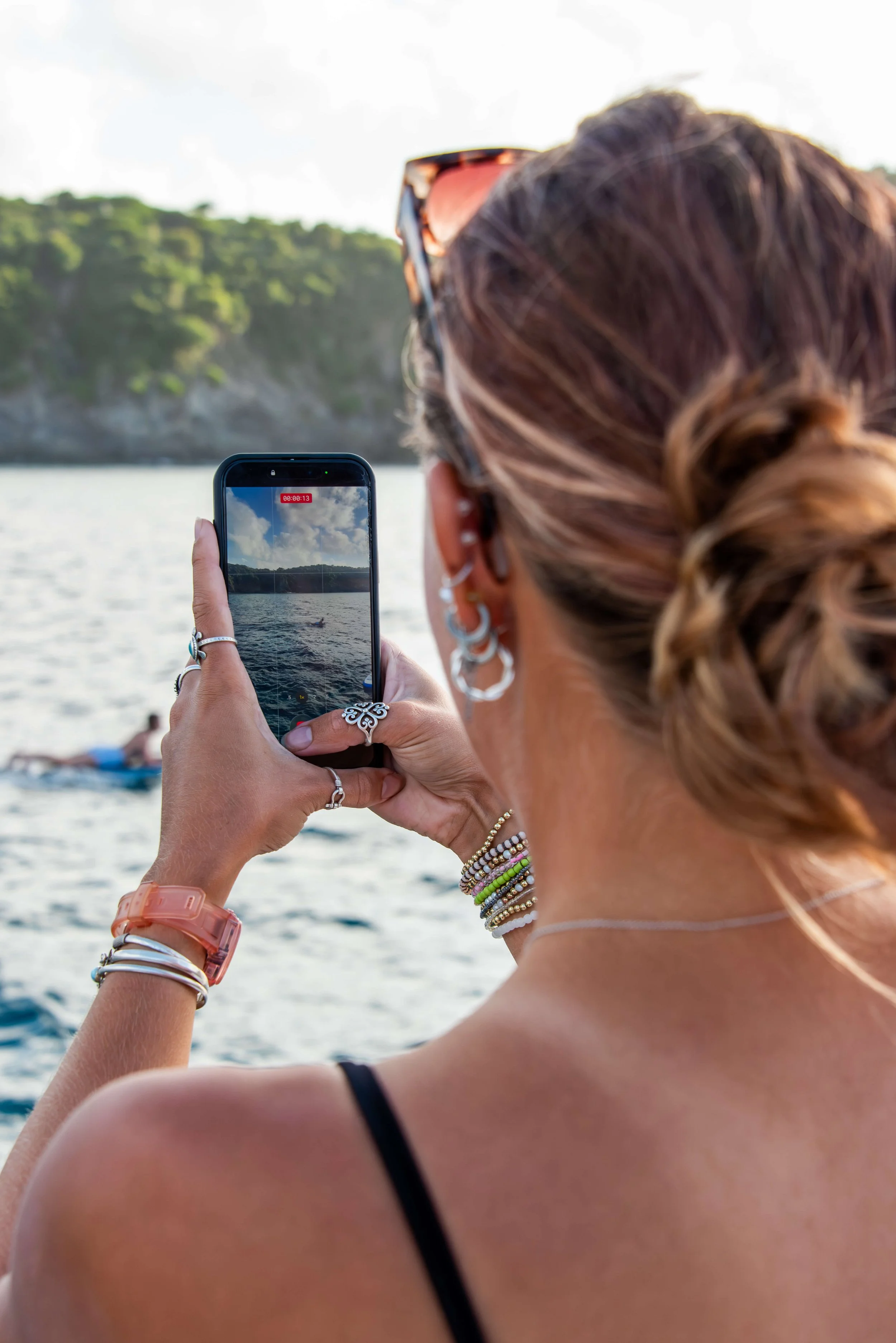 A woman with light brown hair tied in a braid, wearing jewelry and sunglasses, takes a photo of a boat on the water with her phone. In the background, a person is floating on a raft or paddleboard near a shoreline with green trees.
