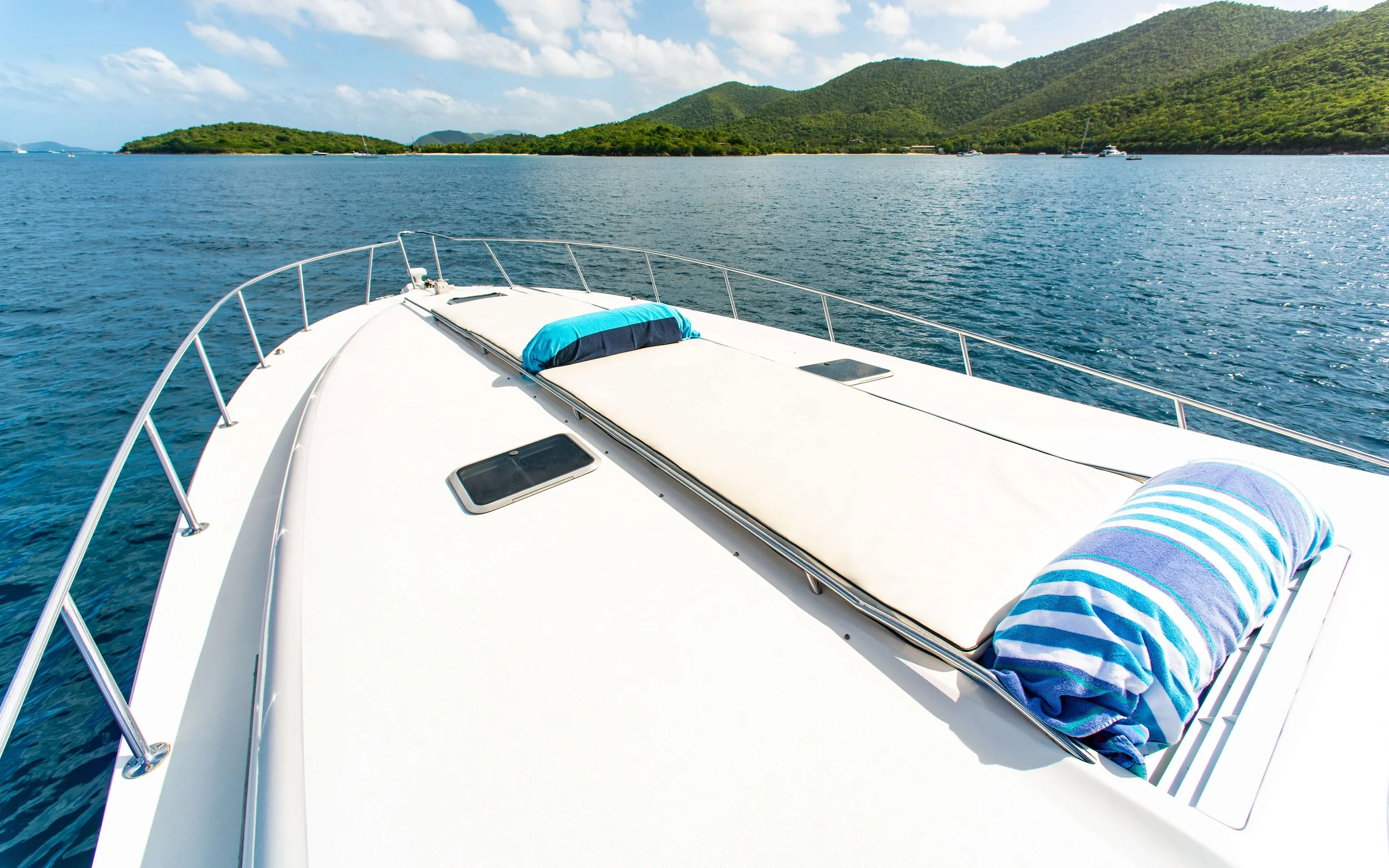 View from the front of a white yacht with two rolled towels, blue and white, on the deck overlooking water and green island with hills in the background.