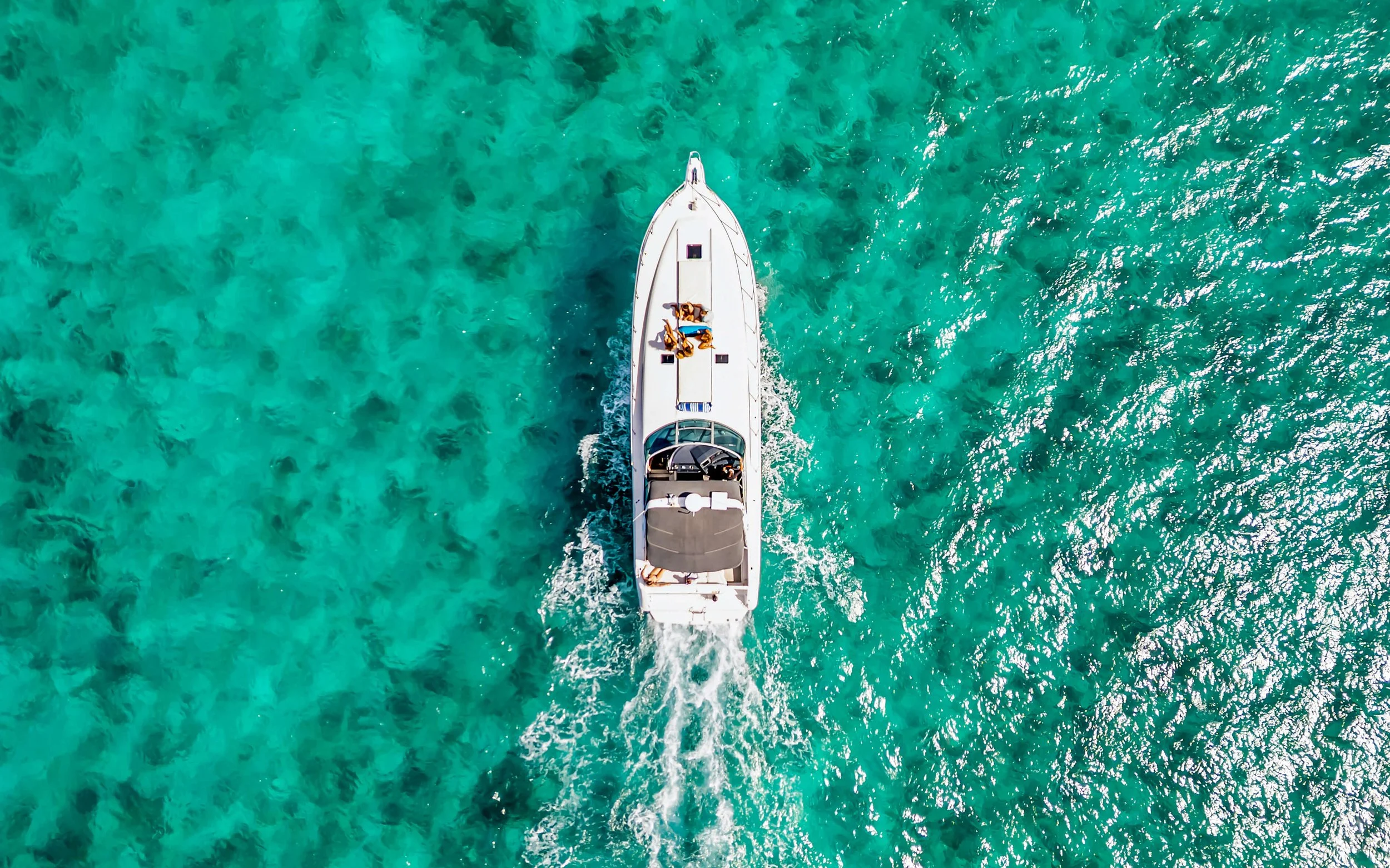Aerial view of a white yacht with three people on the deck, cruising through clear turquoise water.