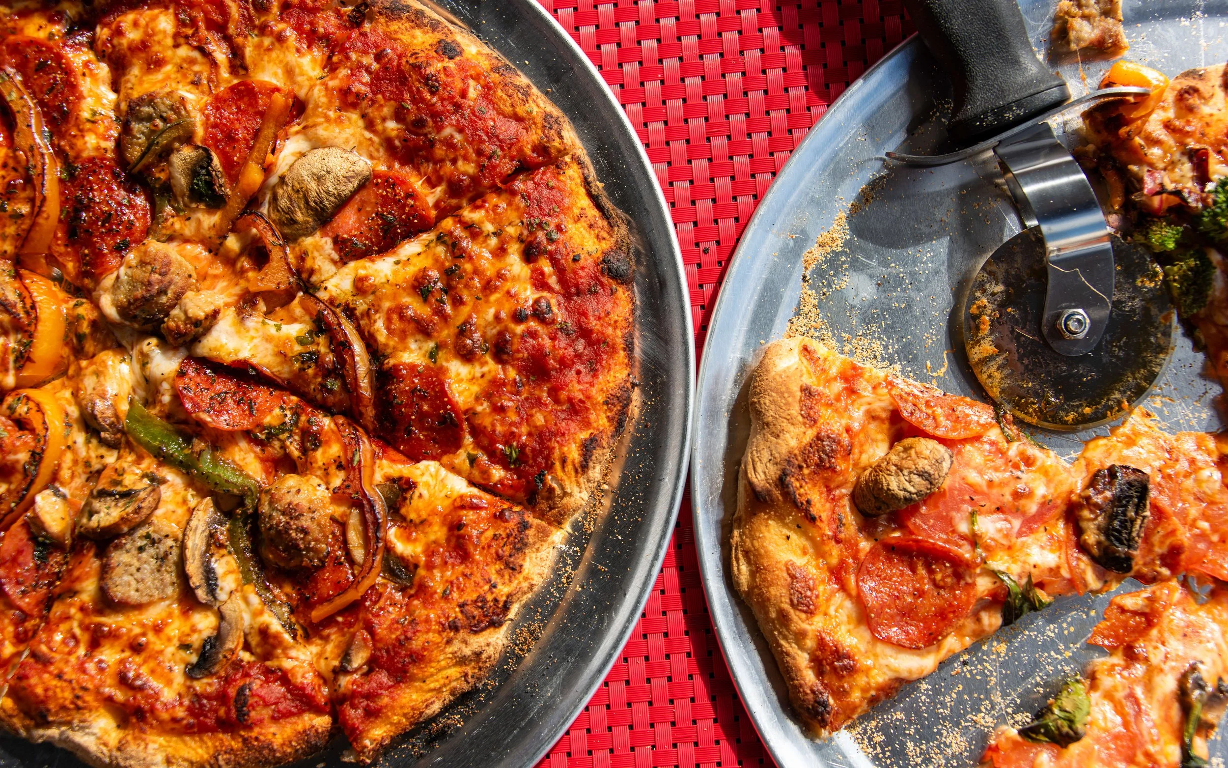 Two pizzas on metal trays, one with toppings including pepperoni, sausage, mushrooms, and green peppers, and the other with pepperoni and a few green peppers, placed on a red woven tablecloth.