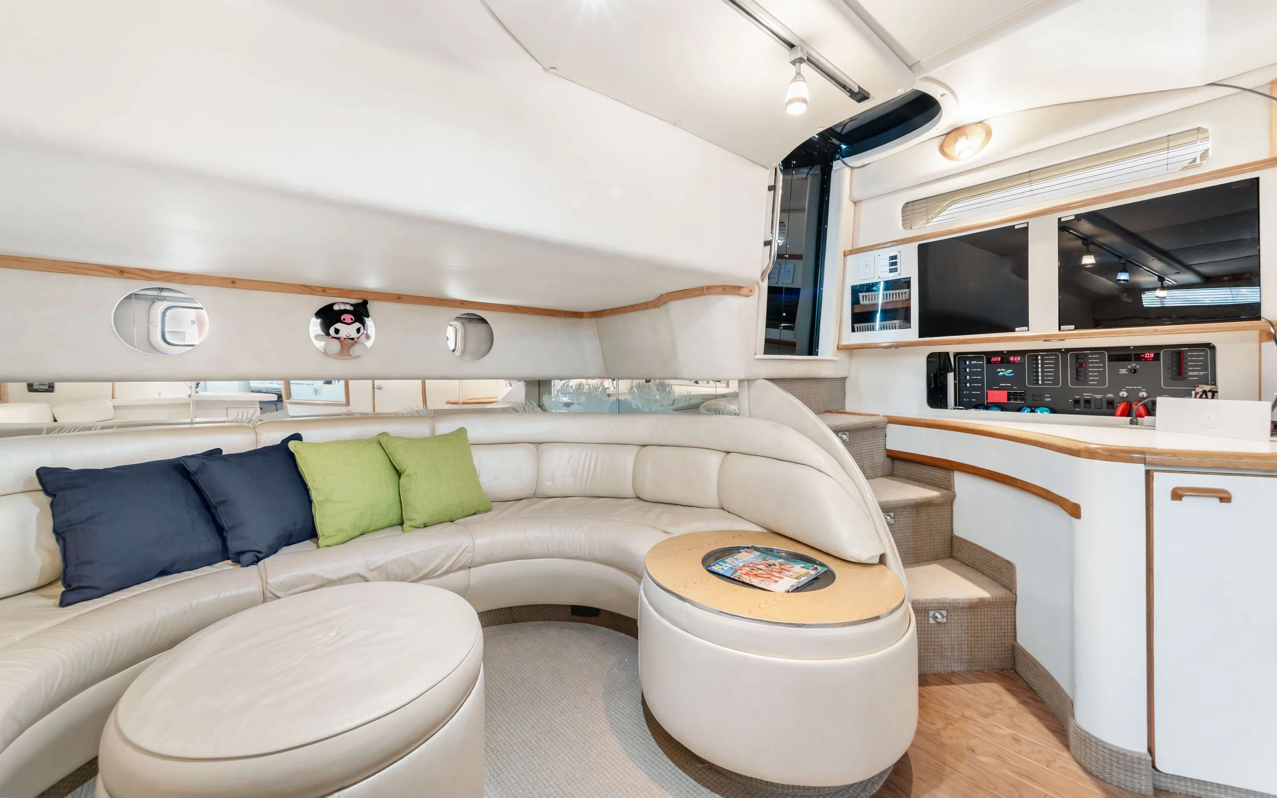 Interior of a yacht with a white curved leather sofa with blue and green pillows, small round ottoman, wooden accents, TV, and control panel, with small portholes in the wall.