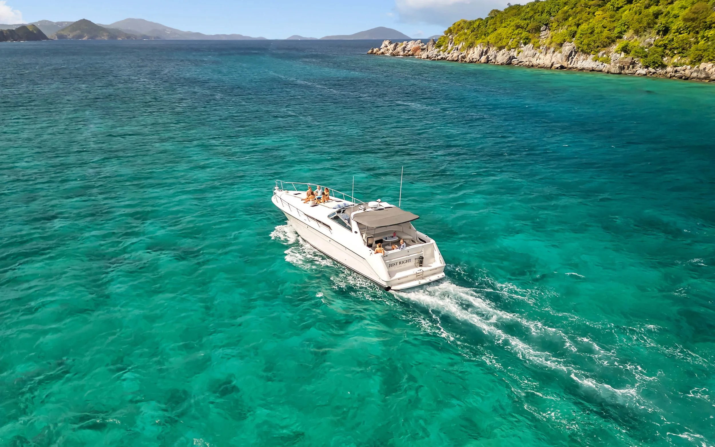 A white motor yacht cruising in clear turquoise waters near a lush green island with rocky shoreline and distant islands in the background.