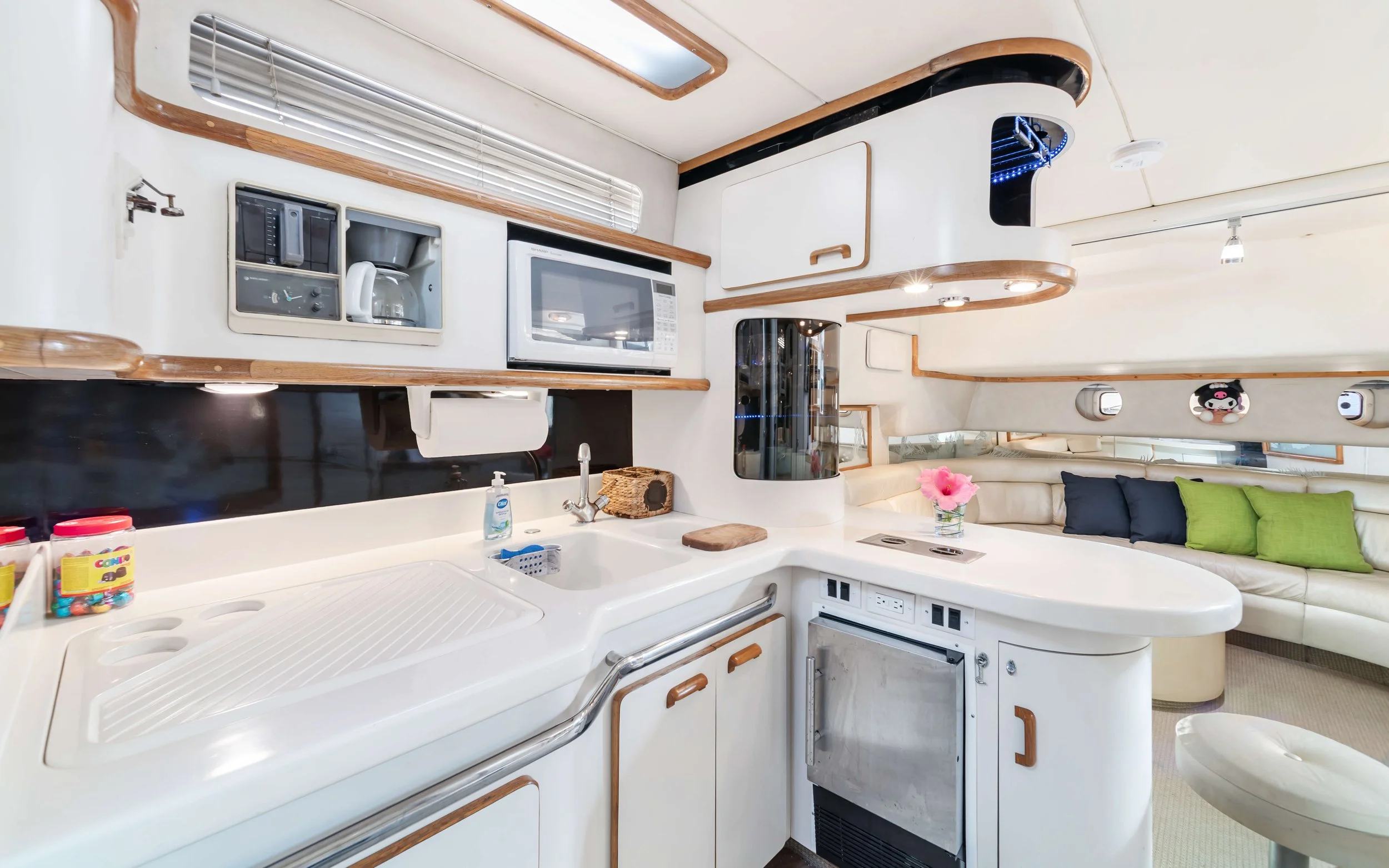 Interior of a compact kitchen and sitting area in a yacht with white cabinets, a microwave, coffee maker, sink, and a curved seating area with pillows and a flower bouquet.