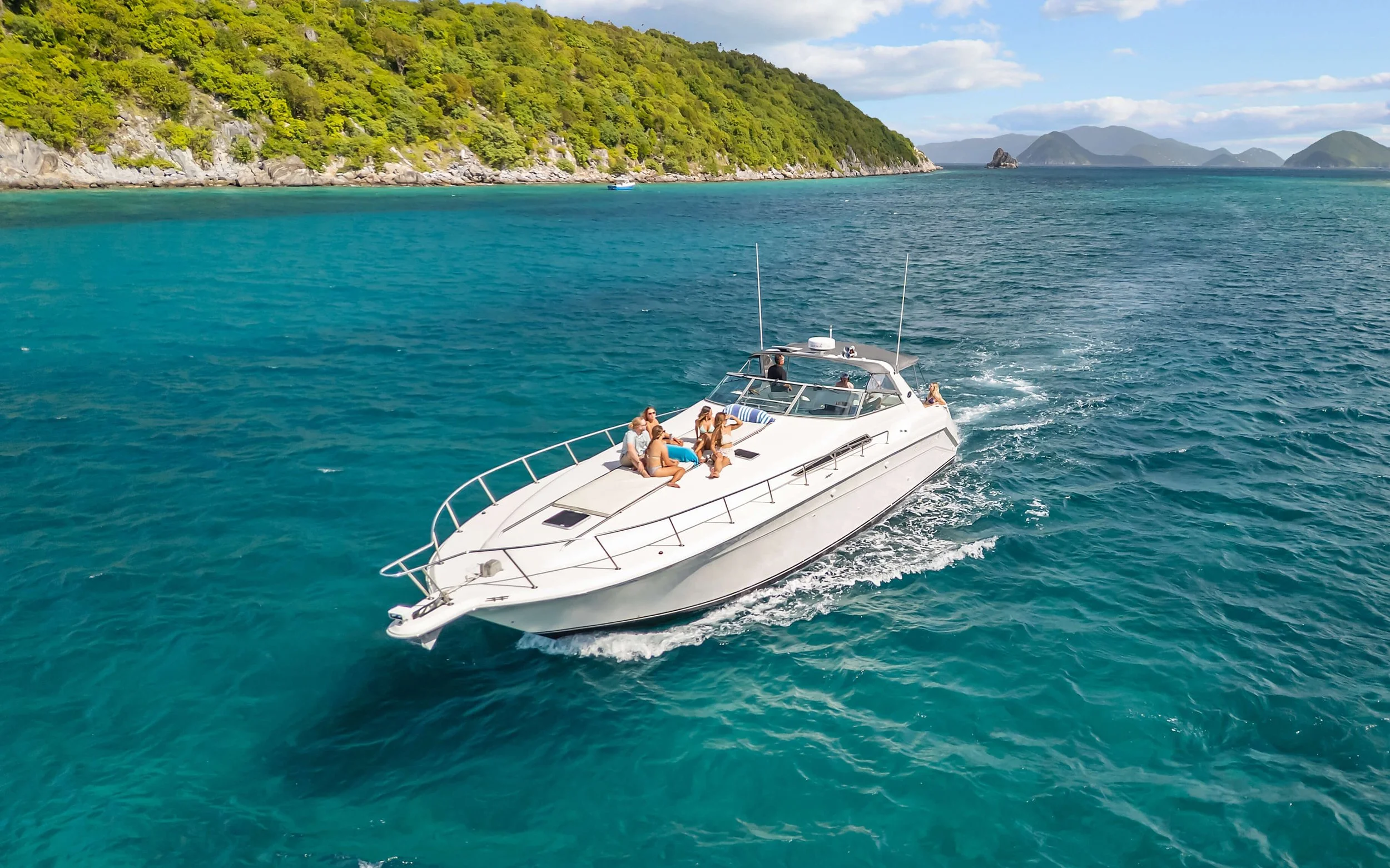 A white motorboat with several people on board sailing on turquoise water near a green, hilly coastline with distant islands.