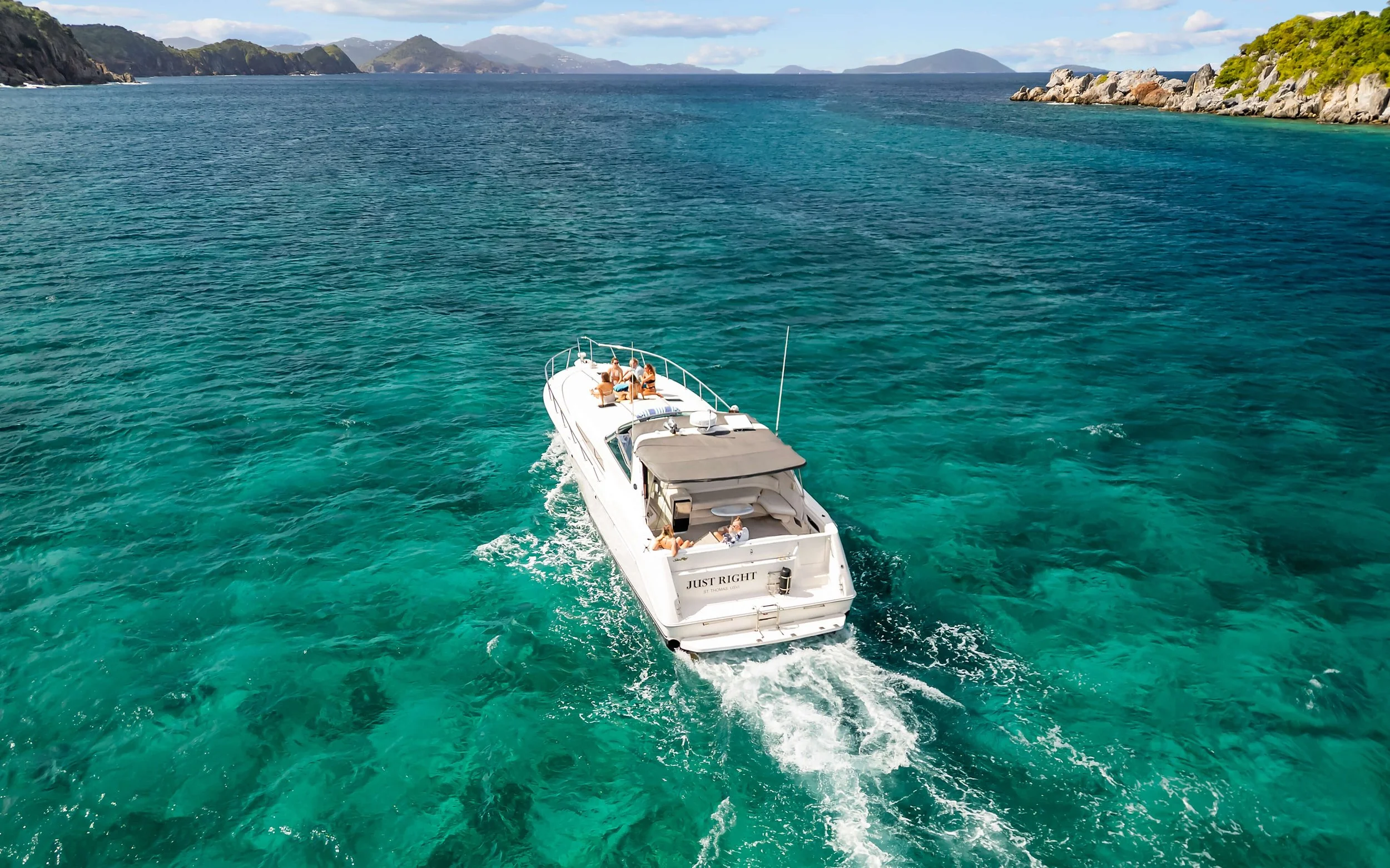 A white yacht named 'Just Right' cruising on clear turquoise water, with a group of people sunbathing and relaxing on deck, surrounded by green islands and hills in the distance.