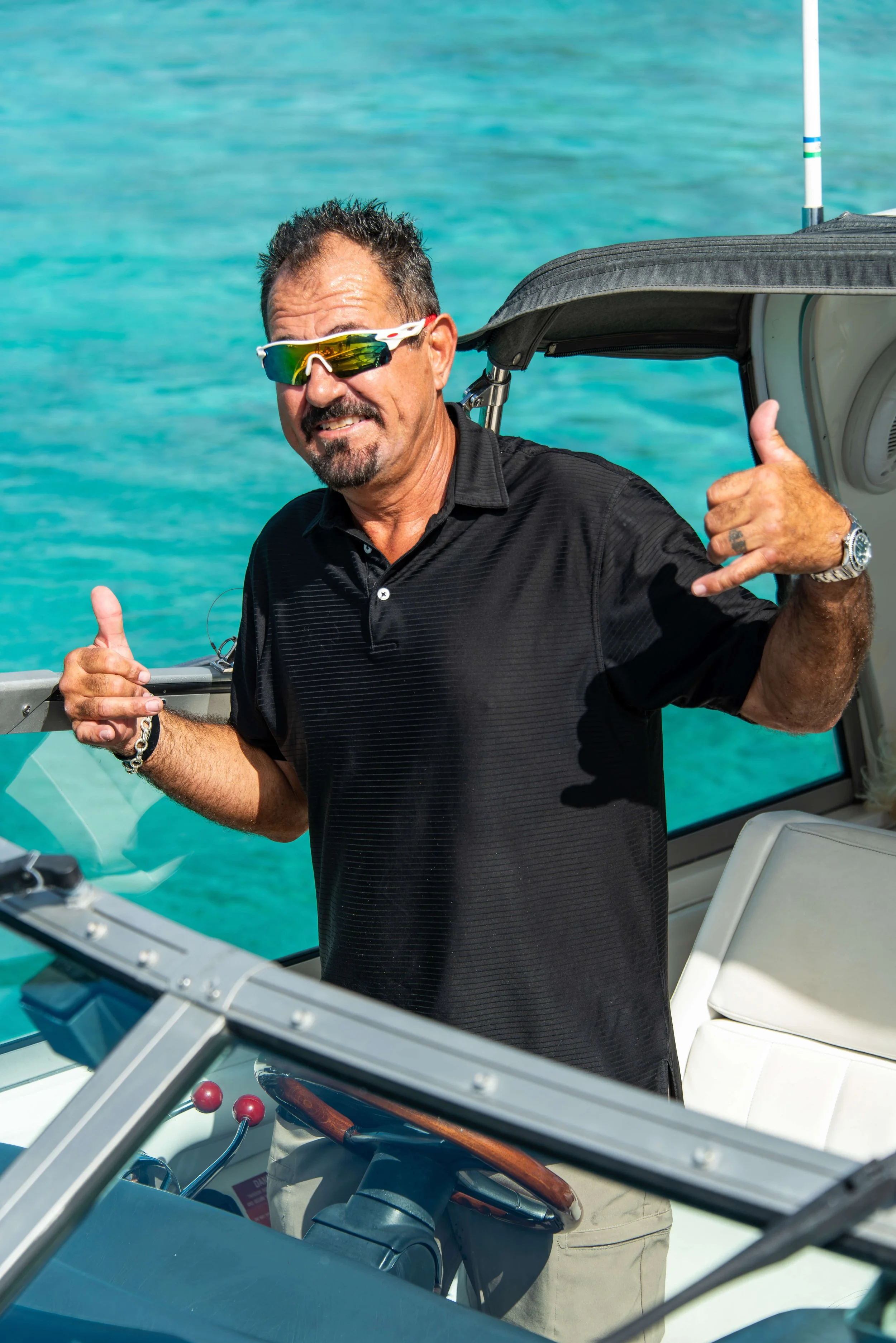A man with sunglasses making a call gesture and a thumbs-up sign on a boat, with ocean water in the background.