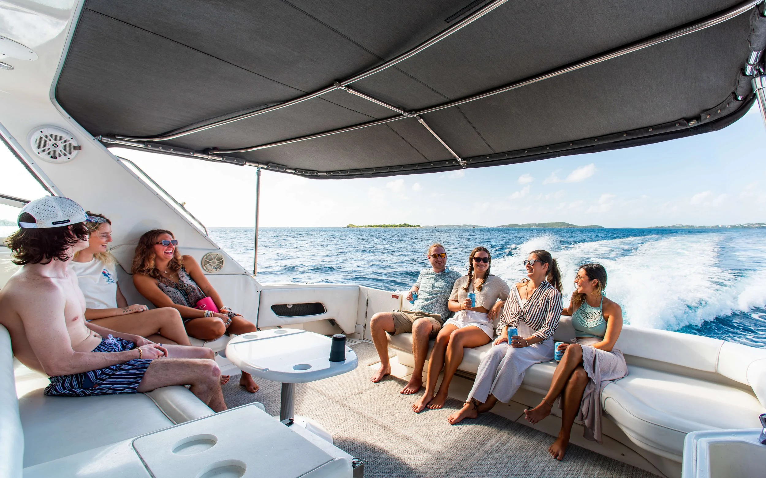 Group of friends enjoying a boat ride on a sunny day, sitting on boat seats with drinks, and the water and sky in the background.
