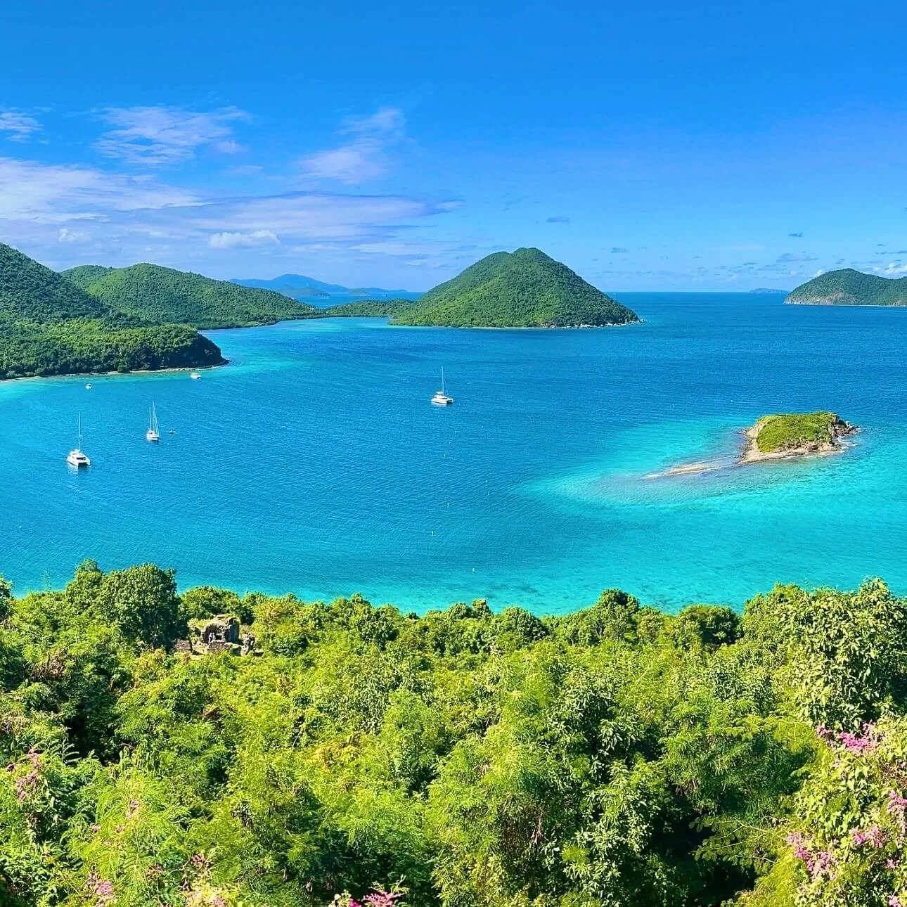 Tropical island view with lush green trees in the foreground, turquoise water with sailboats, and green islands in the distance under a partly cloudy blue sky.