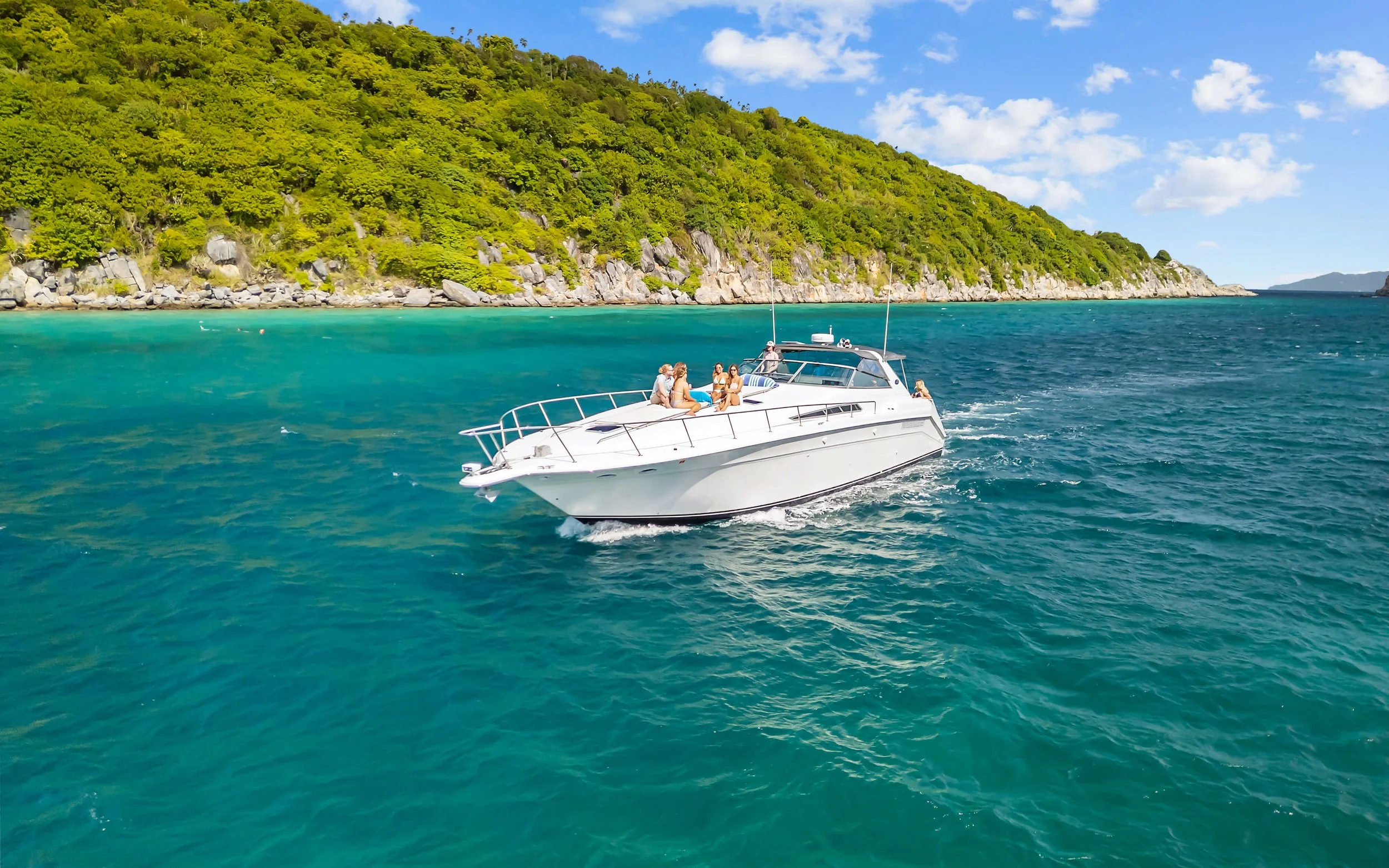 People relaxing on a white motor yacht cruising in clear blue waters near a green, rocky island on a sunny day.