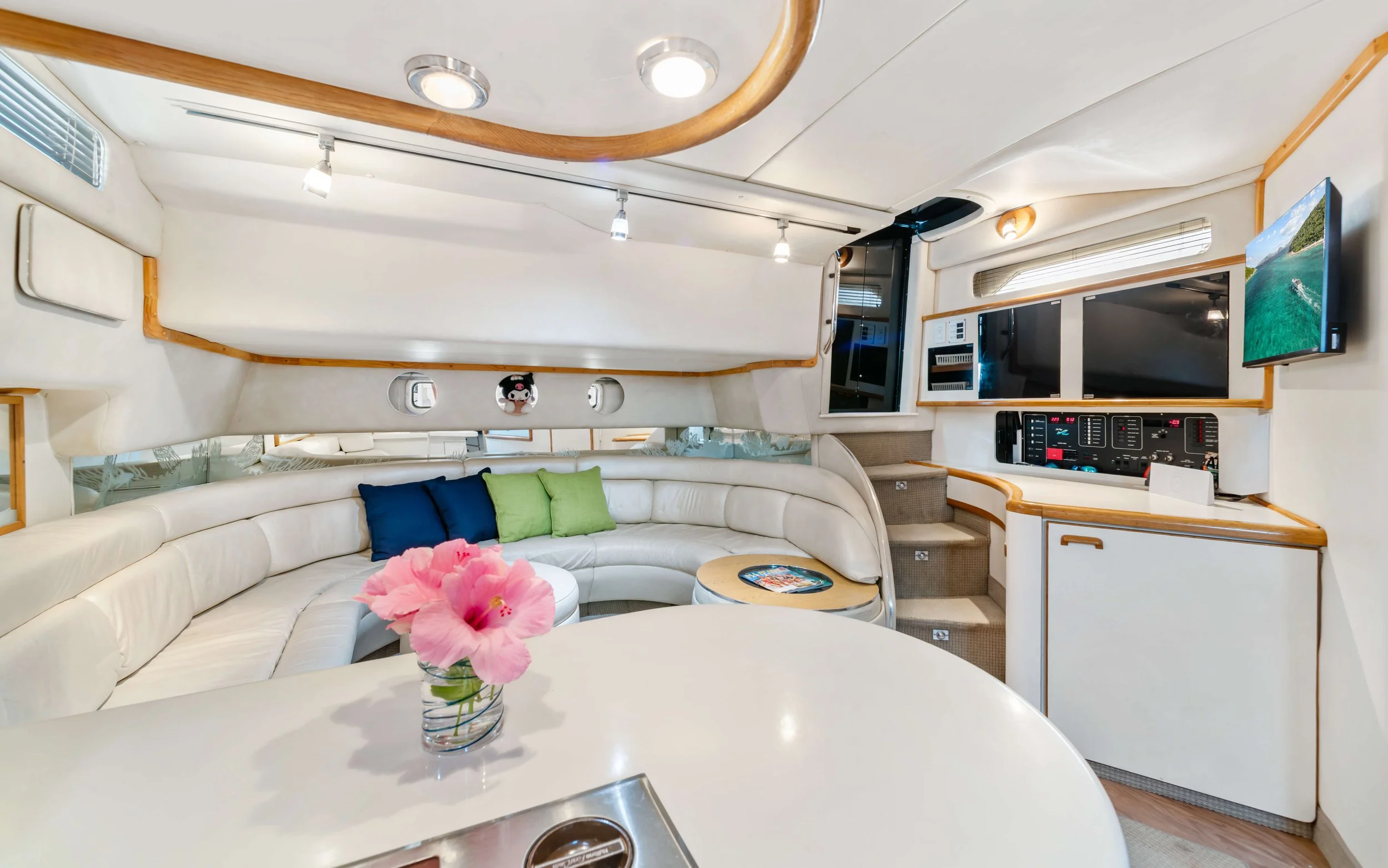 Interior of a yacht with a white curved leather sofa in front of three colorful pillows. A small round table with a magazine and a vase of pink flowers. Multiple screens and control panels are on the wall, with a staircase on the right side. The ceil