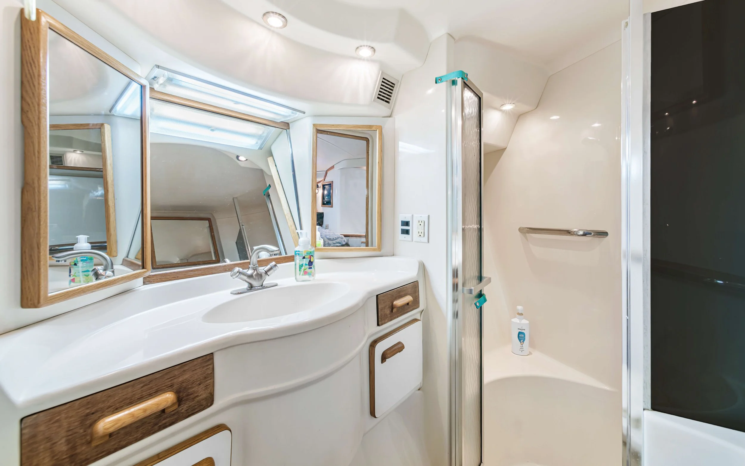 Bathroom with a curved white vanity, three mirrors, and a window above. There is a small bottle of hand soap on the sink, a cleaning spray bottle in one of the mirrors, and a shower to the right with a bottle inside. The bathroom has multiple recesse