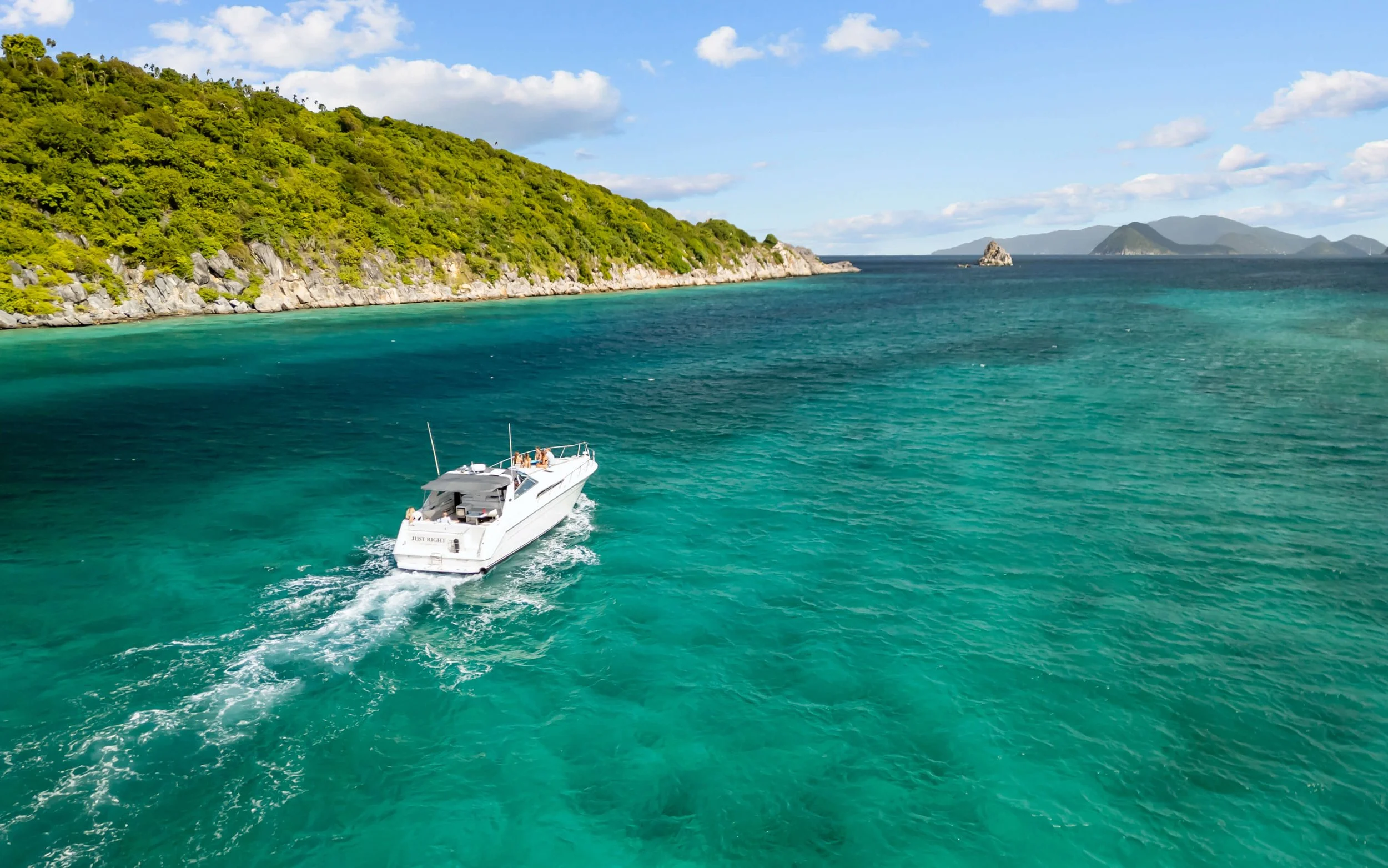 A white yacht sailing through clear turquoise water near a lush, green, rocky coastline with distant islands and a partly cloudy sky.