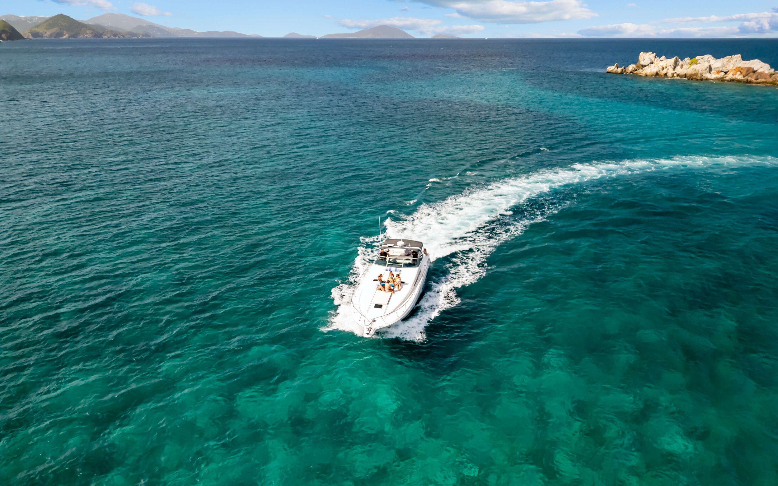 A white motorboat cruising through clear blue ocean water near a rocky shoreline, with distant islands and a partly cloudy sky.