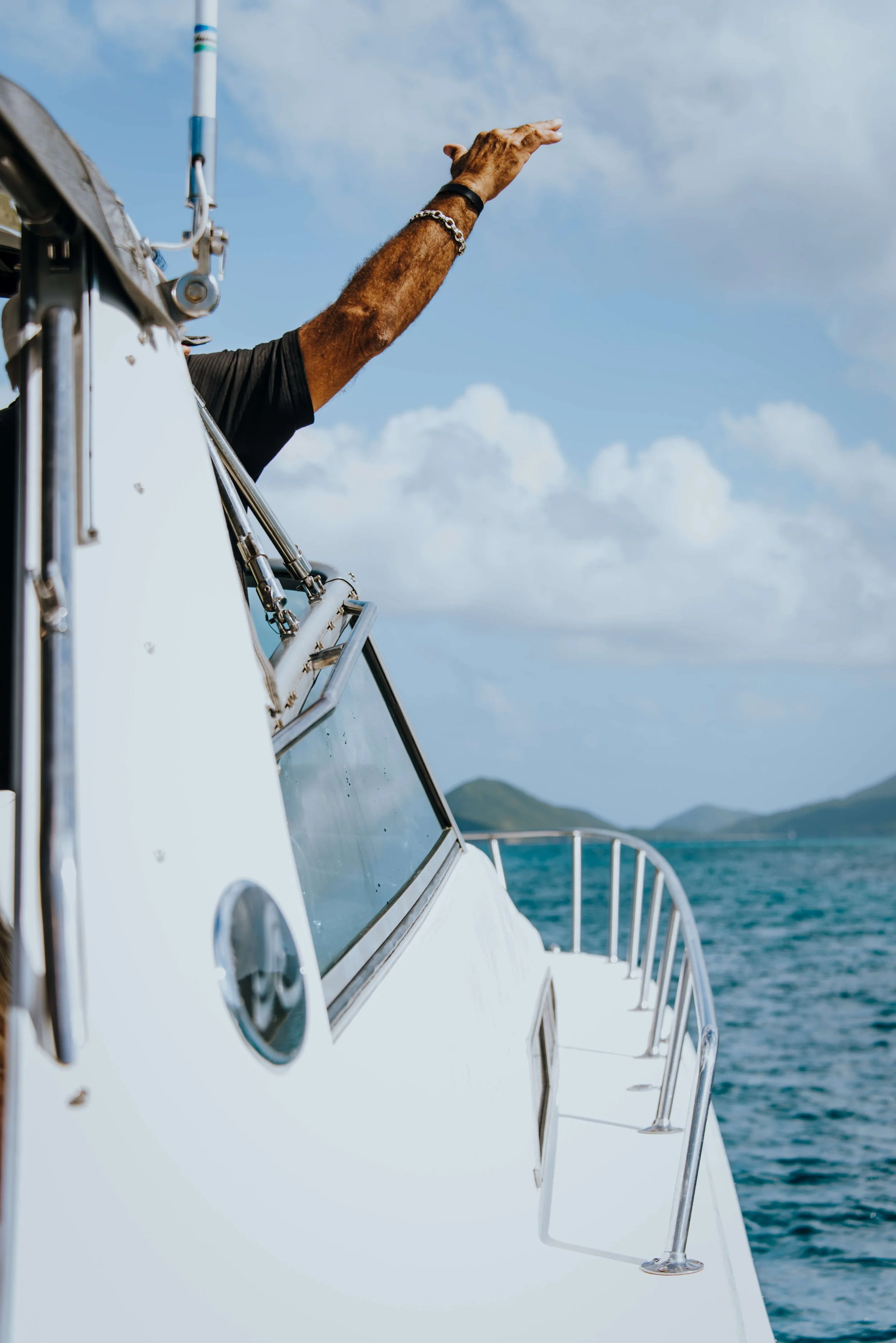 A person on a boat extends their arm outward, waving or reaching towards the sky, with a view of the ocean and distant islands in the background.