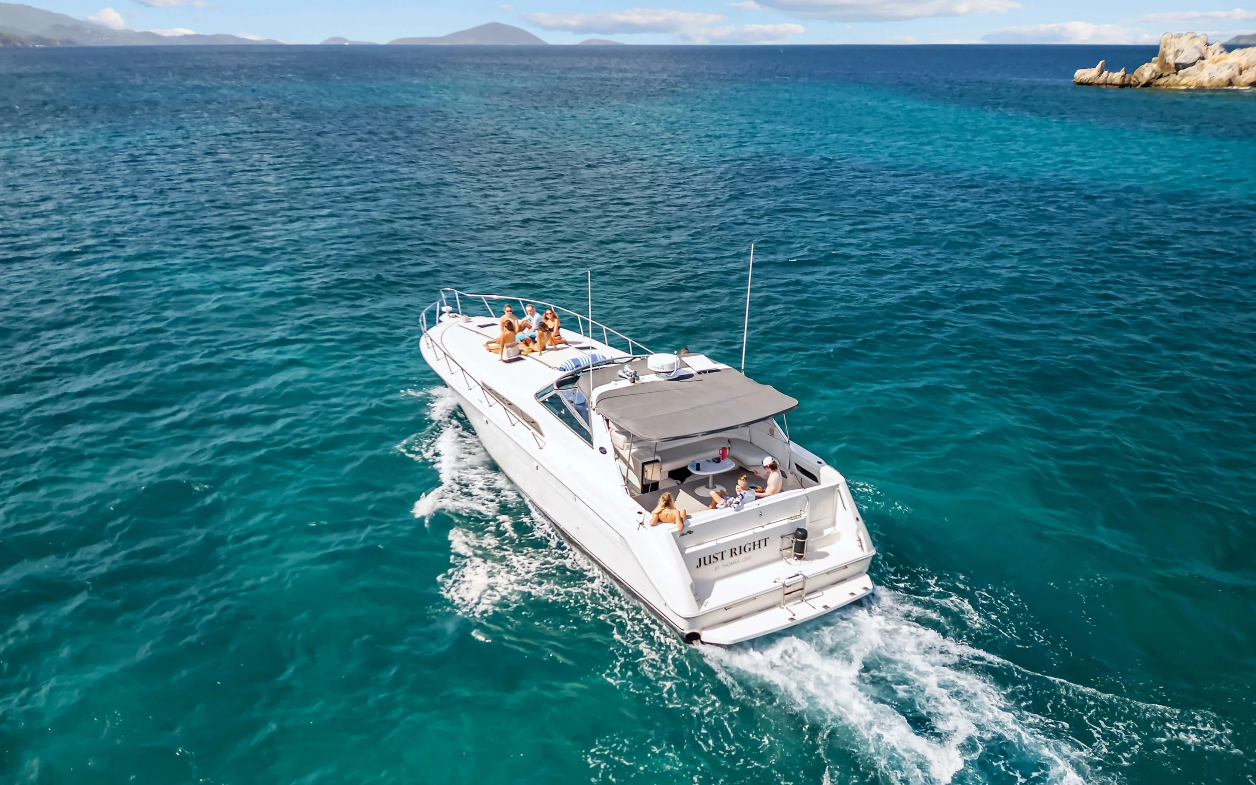 People relaxing and enjoying on a white yacht named 'JUST RIGHT' cruising on clear blue ocean waters with island scenery in the background.