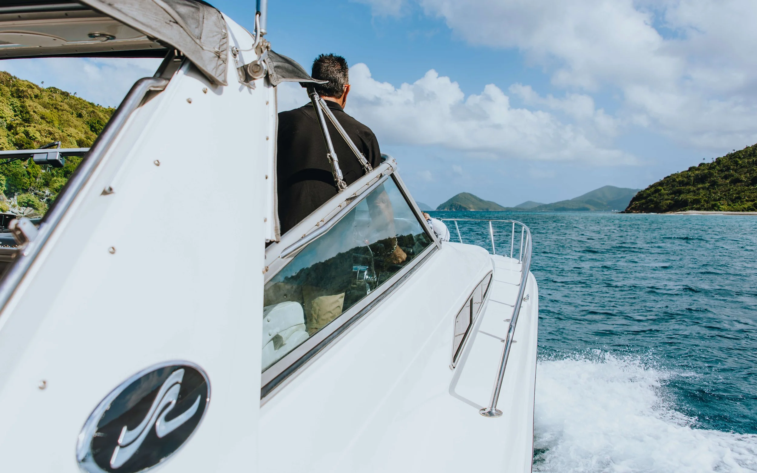 A man steering a white boat on a body of water with green islands and a partly cloudy sky in the background.