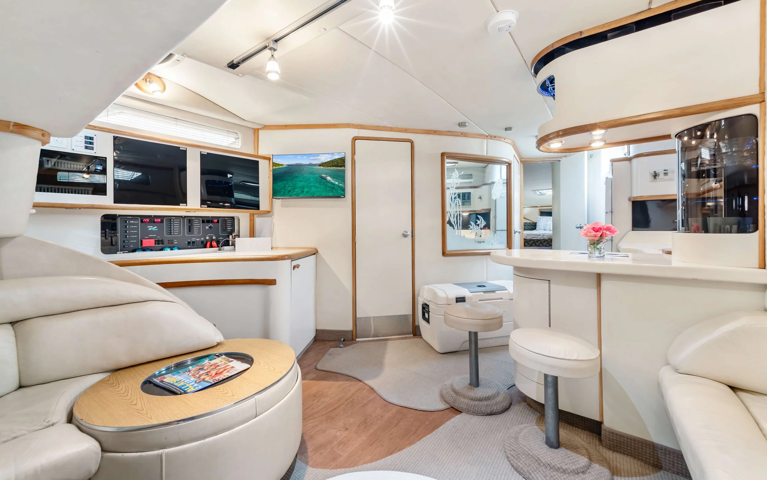 Interior of a yacht living area with cream sofas, a wooden coffee table with a magazine, a mounted television, a control panel, and a small table with pink flowers. There are compartments, mirrors, and a door leading to another room.