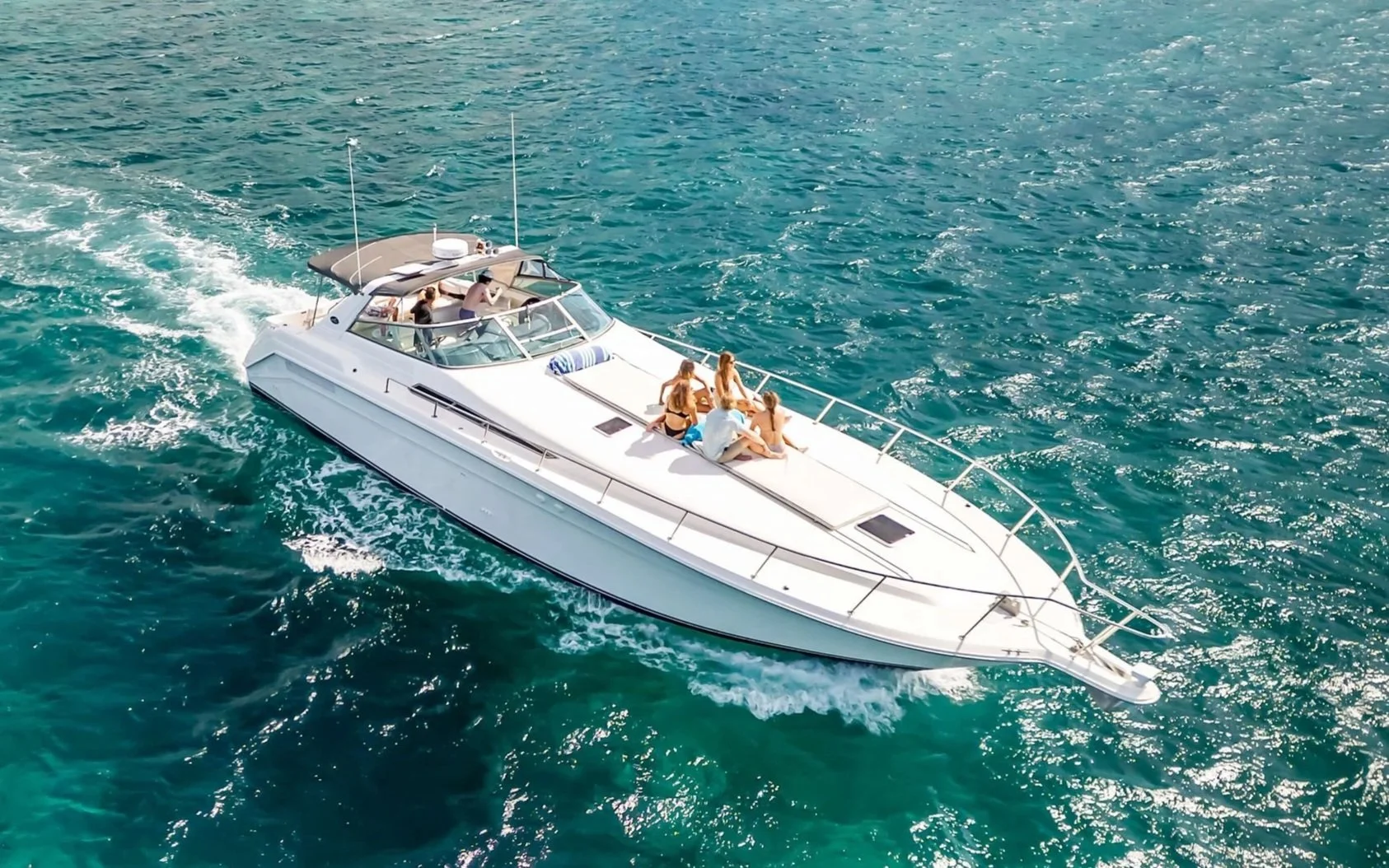 A white yacht with four people lounging on the deck, sailing in clear blue ocean waters.