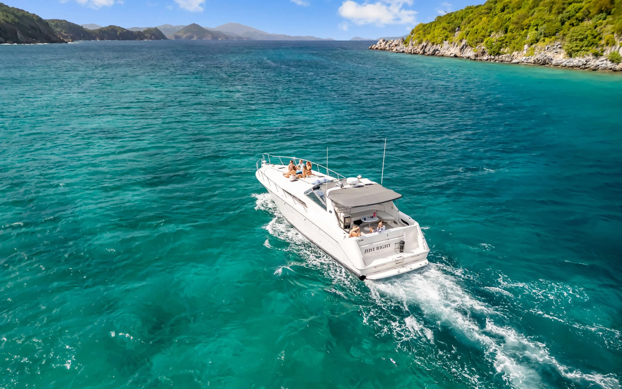 A white yacht named "Just Right" traveling on turquoise waters near rocky and green shoreline with mountains in the background.