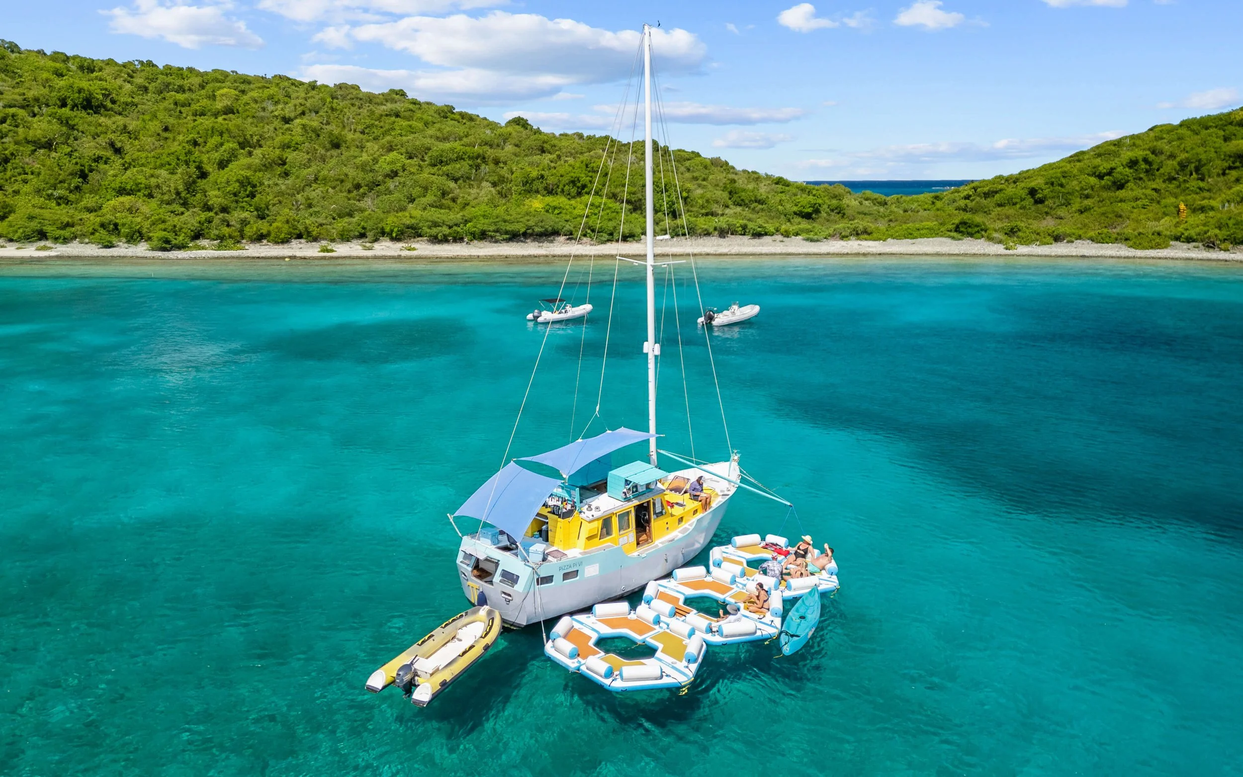 A sailboat anchored in turquoise waters near a green island with two inflatable floating platforms with people relaxing and sunbathing.