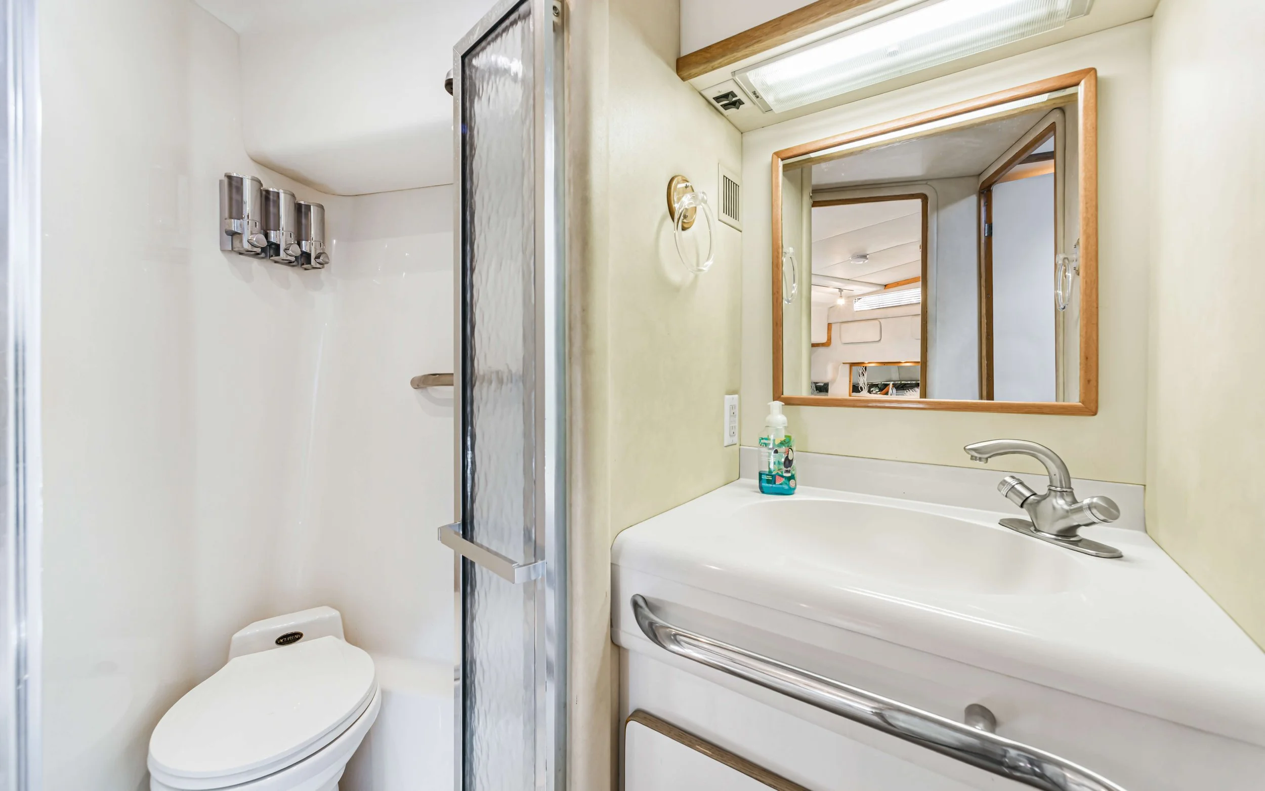 Bathroom with a small sink, mirror, soap dispenser, and a shower with a glass door, toilet, and white walls.