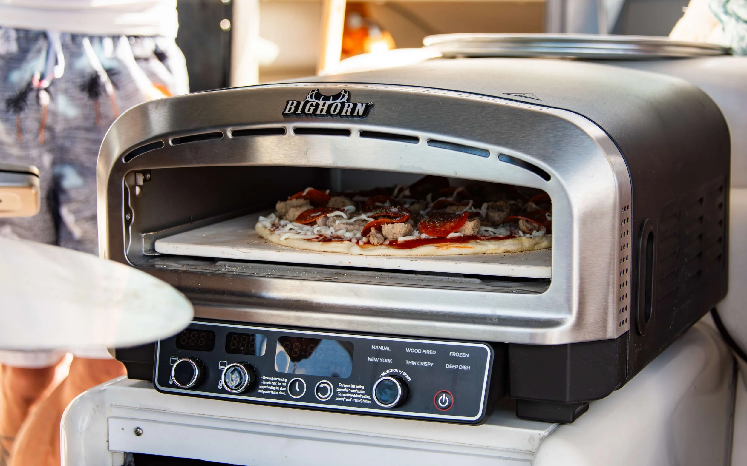 A pizza is being baked in an industrial pizza oven, branded Bighorn, with toppings visible on the pizza crust.