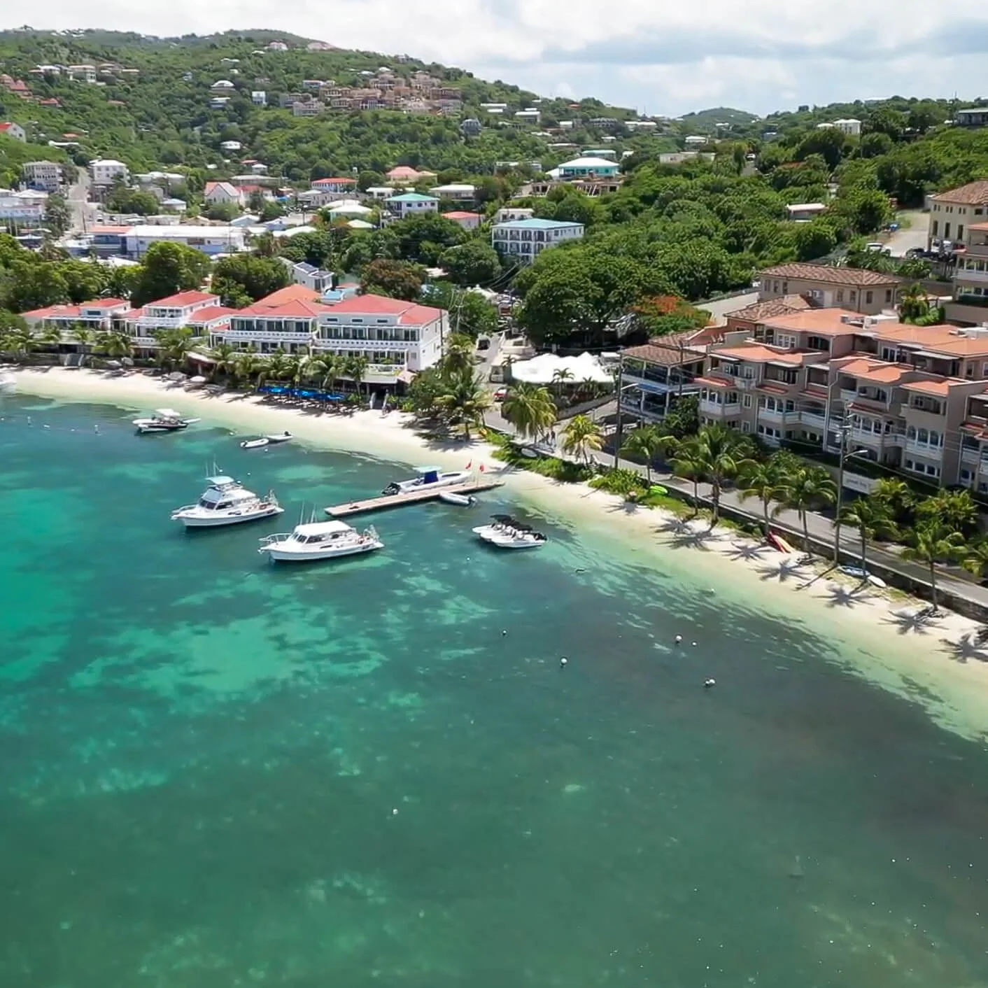An aerial view of a tropical coastal town with colorful buildings, palm trees, and boats docked along a sandy beach, with lush green hills in the background.
