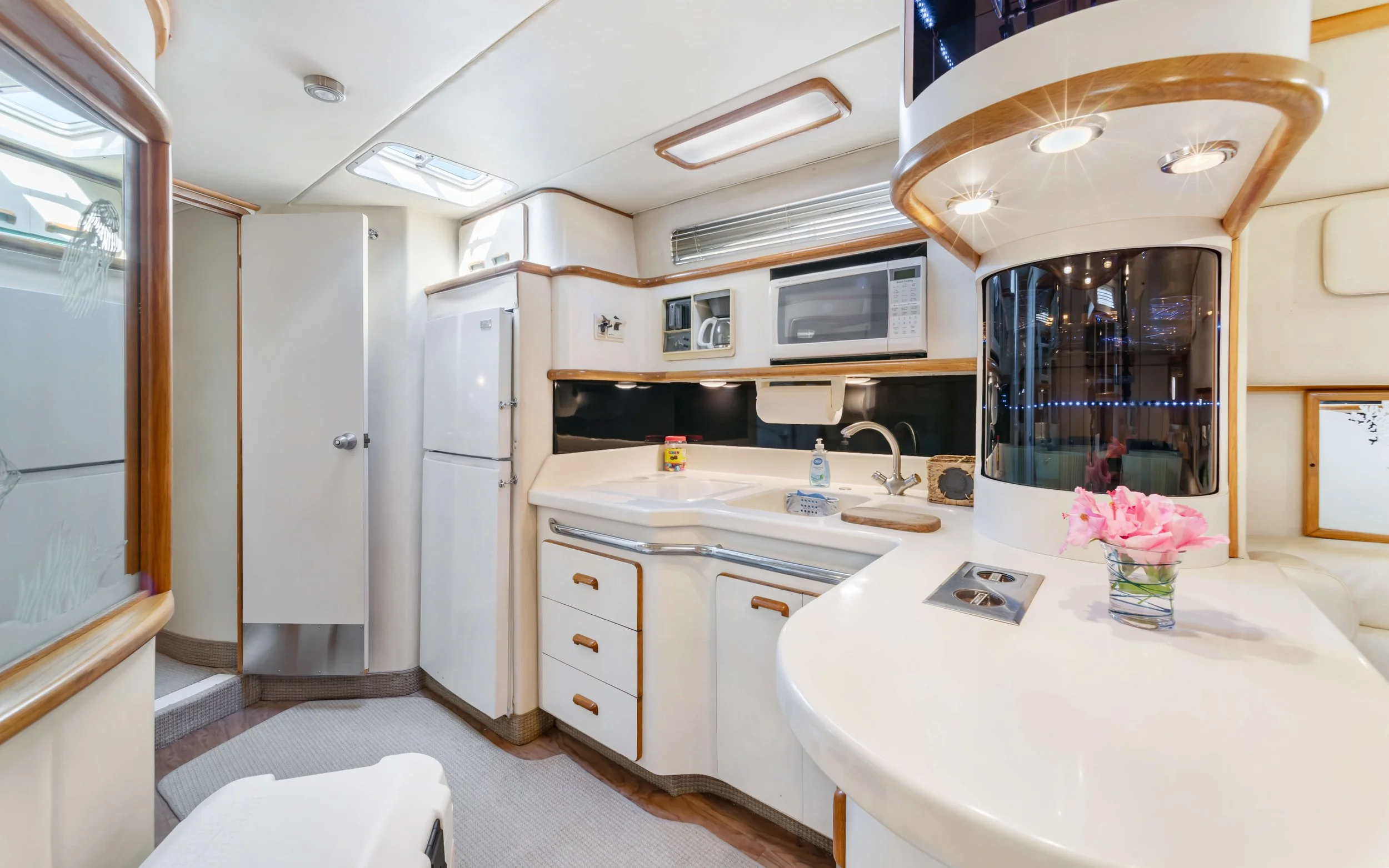 Interior view of a compact boat or RV kitchen with white cabinets, a sink, microwave, and refrigerator. Pink flowers in a glass vase sit on the counter.