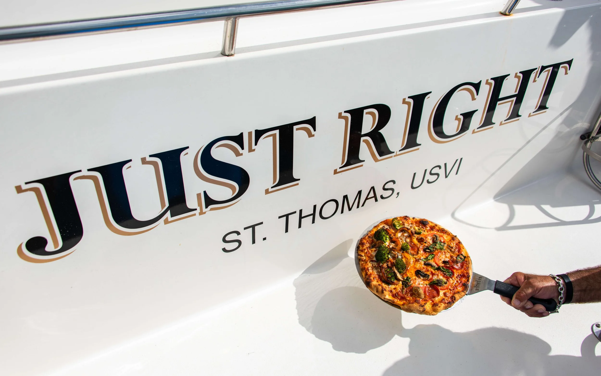 A person holds a pizza with various toppings, including green peppers, mushrooms, and sausage, near a white boat with the text 'Jusit RIGH' and 'St. Thomas, USVI'.