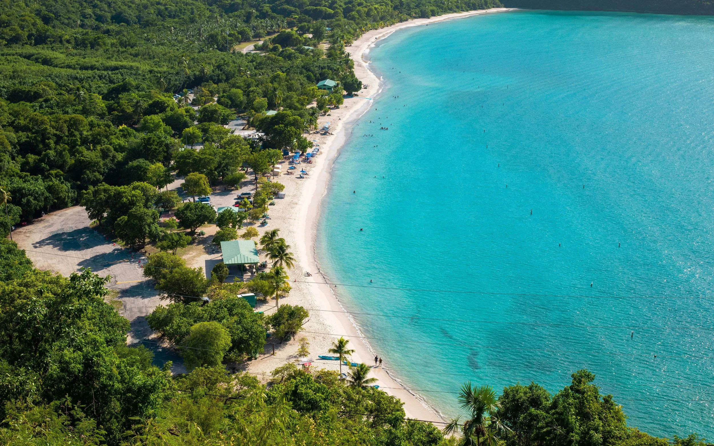 Aerial view of a tropical beach with turquoise water, white sandy shoreline, lush green trees, and small buildings.